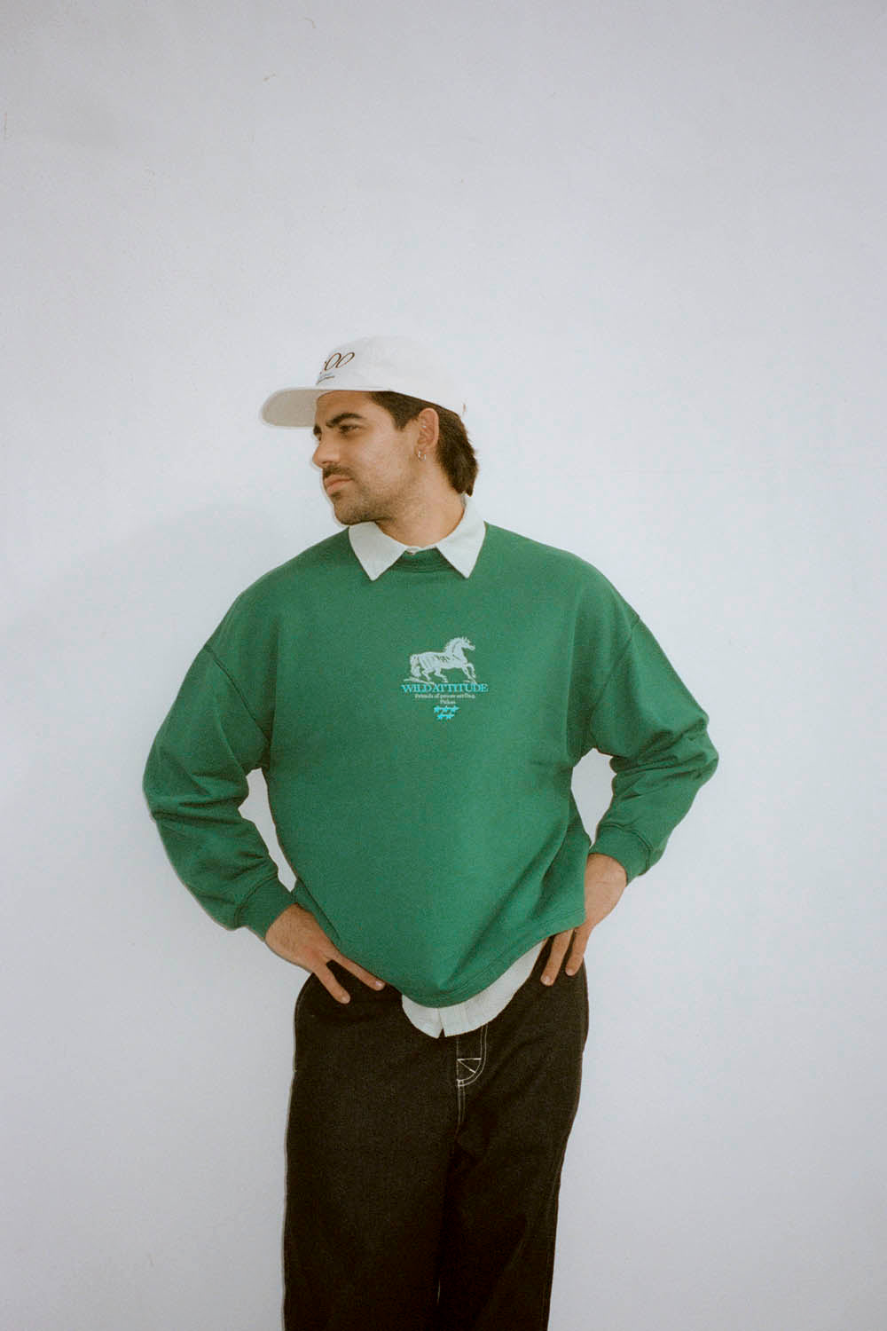Man wearing a green sweatshirt with a logo, white cap, and dark pants on a plain background