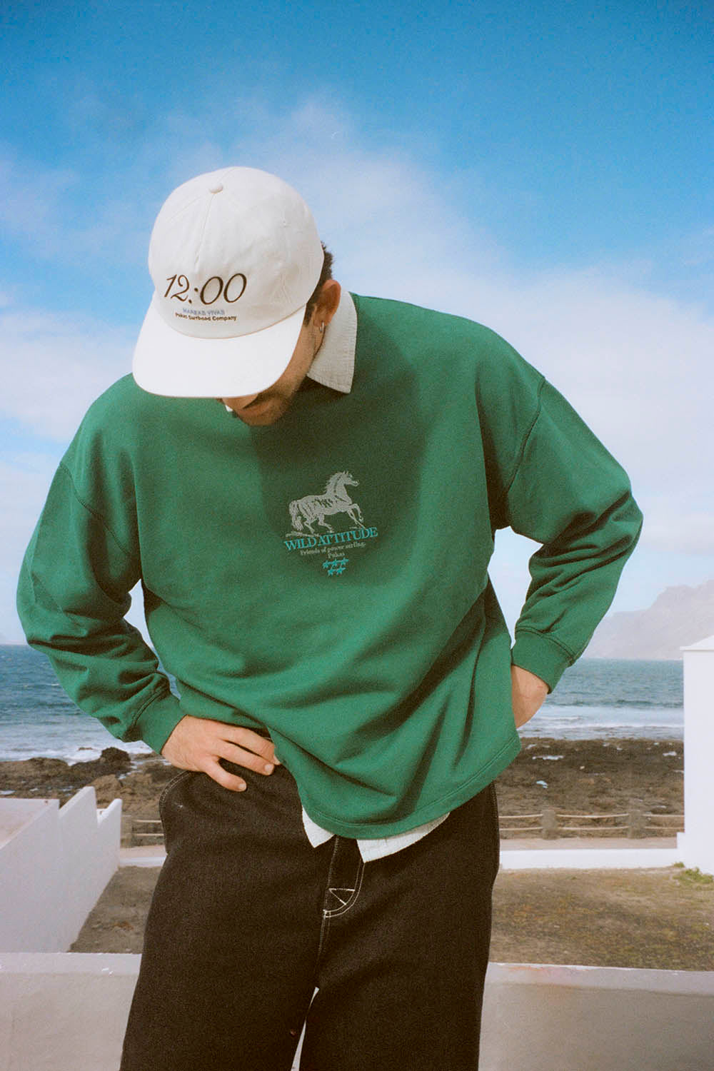 Person wearing a green sweatshirt with a logo and a white cap, standing by the ocean.