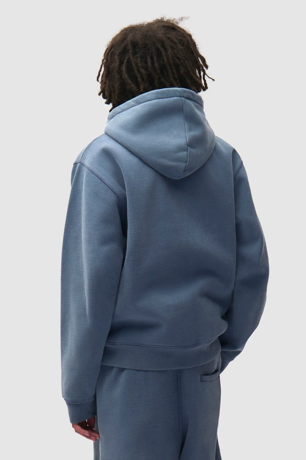 A person shown from the back wearing a dusty blue pullover hoodie and matching sweatpants against a light gray studio background.