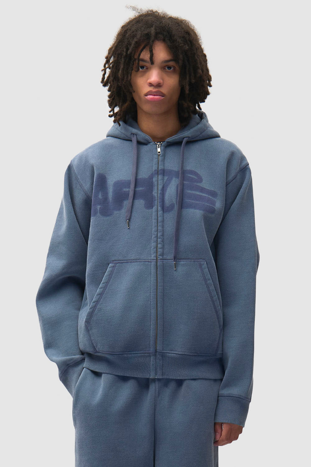A model wearing a slate blue zip-up hoodie with a tonal dark blue 'ART' graphic across the chest, paired with matching blue sweatpants, against a light gray background.