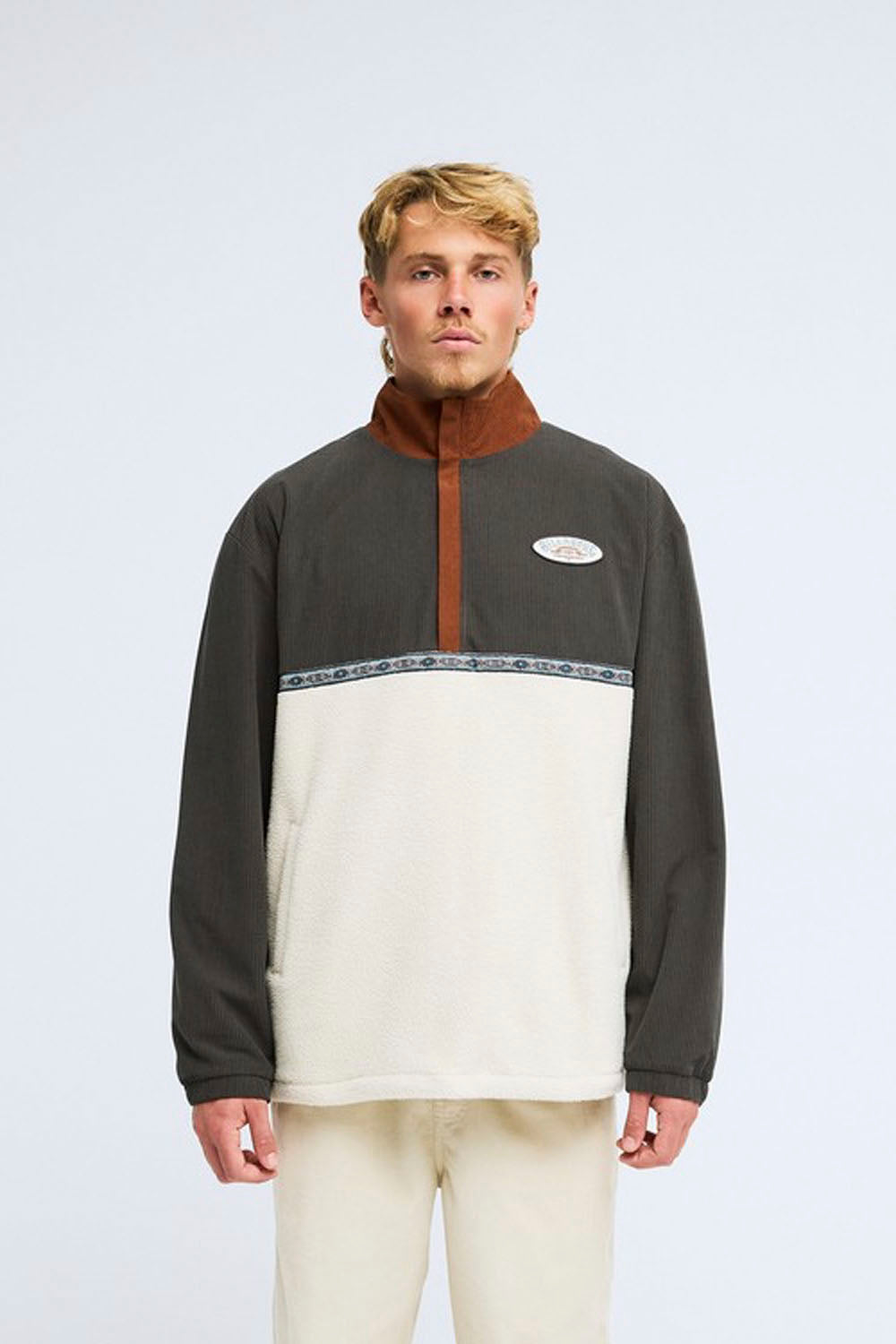 A man wearing a color-blocked quarter-zip fleece pullover with a dark gray upper section, a brown collar and zipper placket, and an off-white lower section, paired with off-white pants, against a light gray studio background.