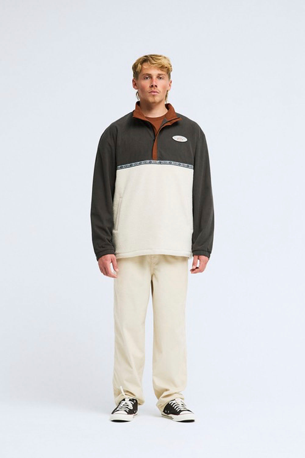 A man stands against a light gray studio background wearing a color-blocked quarter-zip fleece pullover with a dark gray upper section, a cream lower section, and a brown collar, paired with cream sweatpants and black-and-white sneakers.