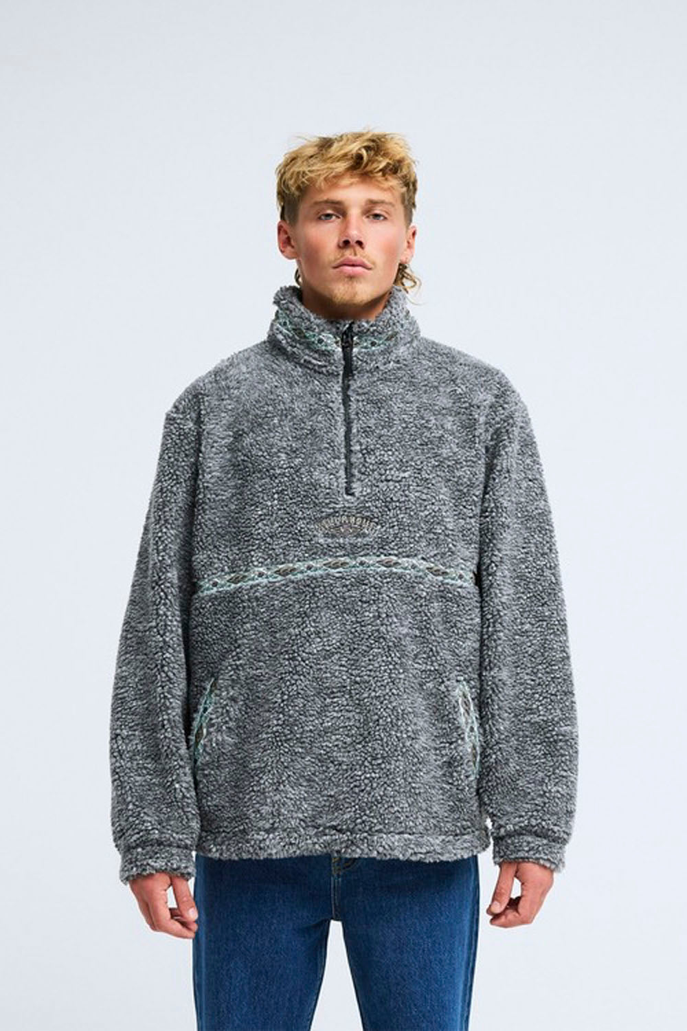 A man wearing a gray marled quarter-zip fleece pullover sweater with a horizontal knit stripe detail across the chest, paired with blue jeans, against a light gray studio background.