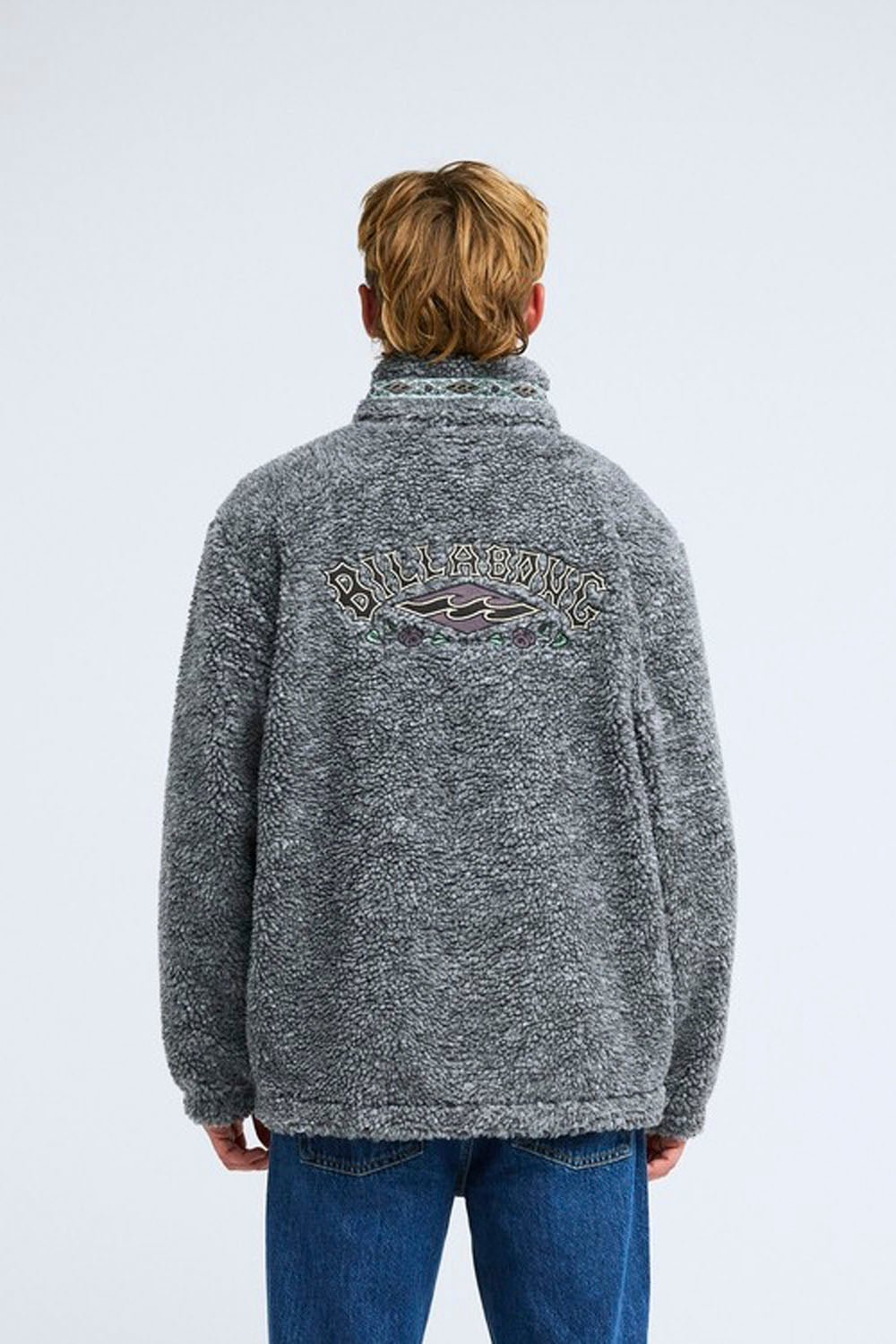 A man viewed from behind wearing a heather gray fleece quarter-zip jacket with a large Billabong logo on the back, paired with blue jeans, against a light gray studio background.