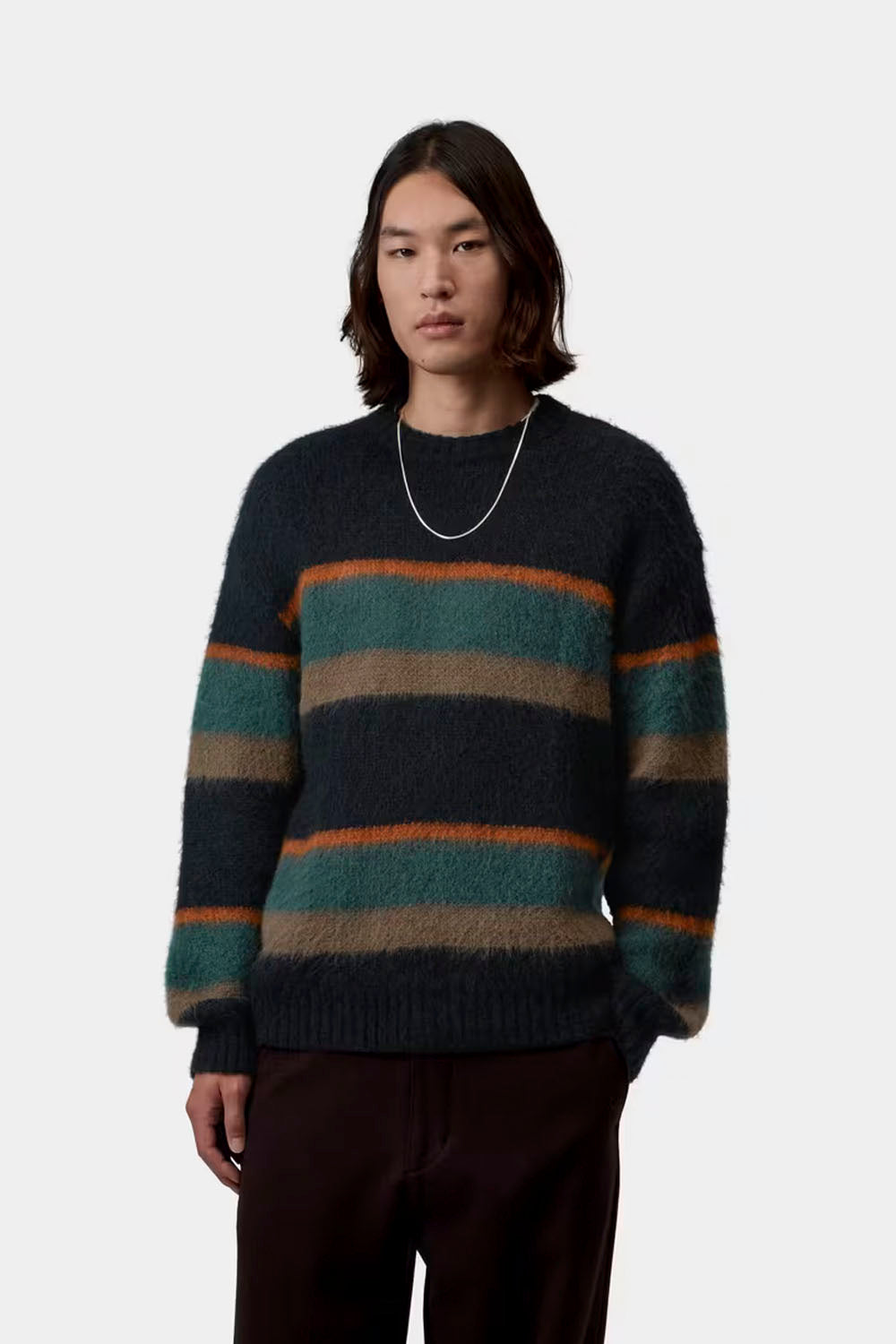 A model wearing a fuzzy, long-sleeve crewneck sweater with horizontal stripes in dark navy, teal green, brown, and orange, paired with dark brown trousers and a silver chain necklace, against a light gray background.