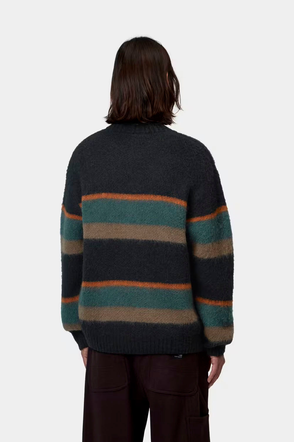 A person shown from the back wearing a fuzzy knit sweater with horizontal stripes in dark gray, teal green, brown, and rust orange, paired with dark brown pants, against a light gray studio background.