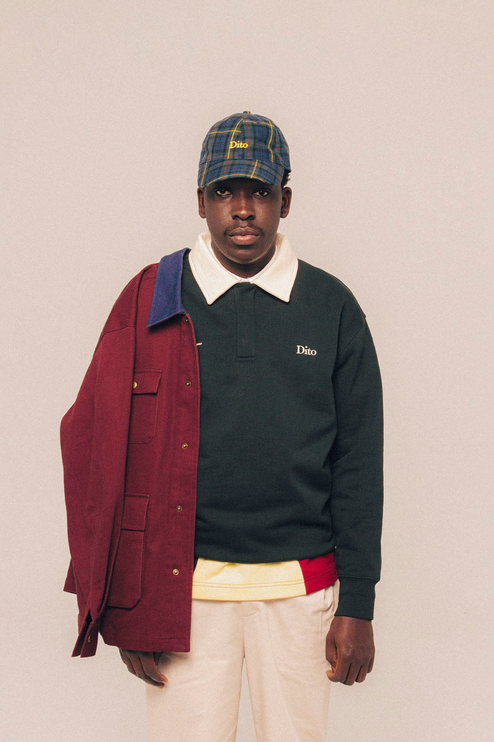 A model wearing a dark green Dito long-sleeve polo sweater with a white collar, a burgundy overshirt draped over one shoulder, cream trousers, and a blue plaid cap, set against a solid light beige background.
