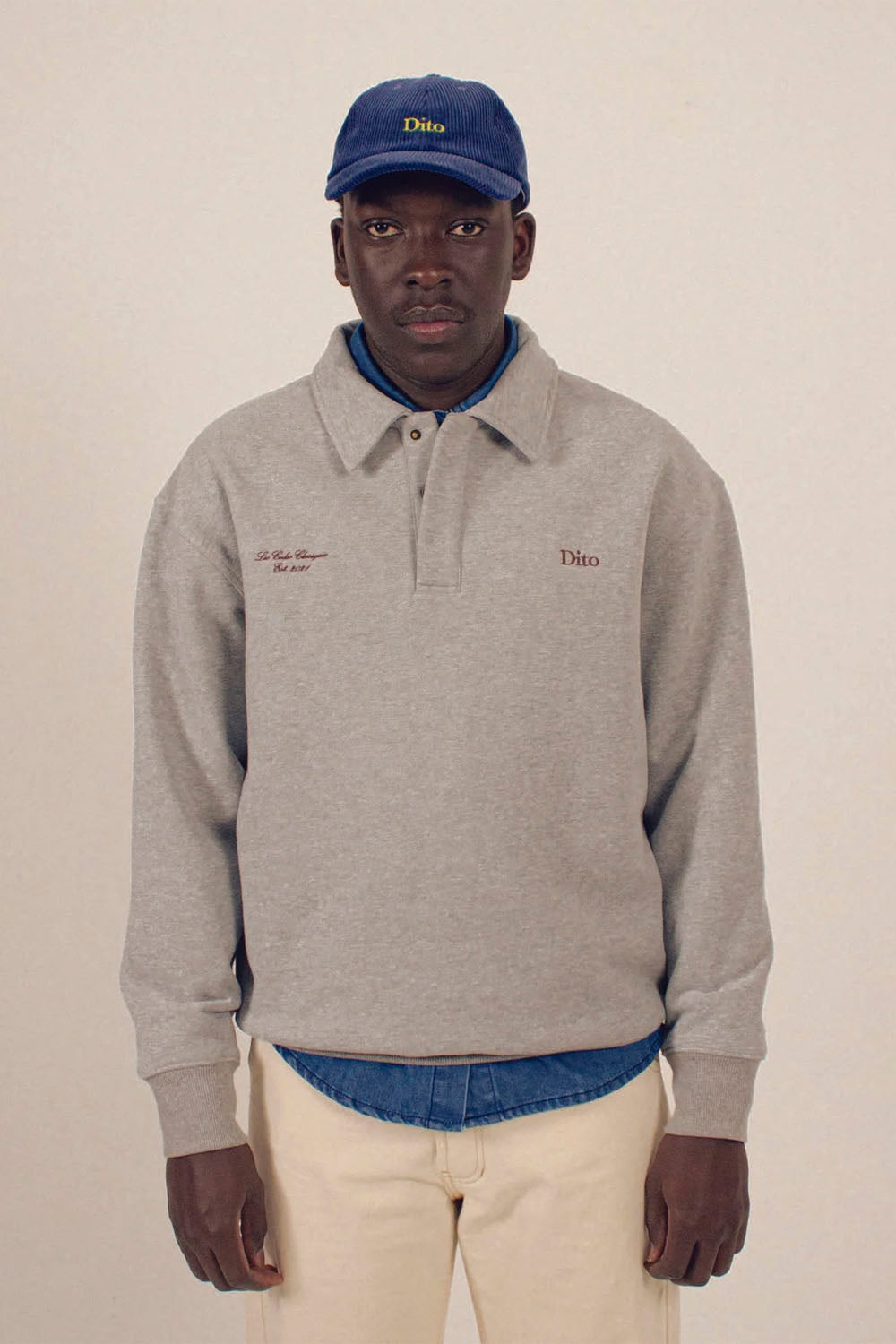 A man wearing a light gray long-sleeve polo sweatshirt with 'Dito' embroidered on the chest, paired with a blue cap, blue shirt underneath, and beige pants, against a plain beige background.