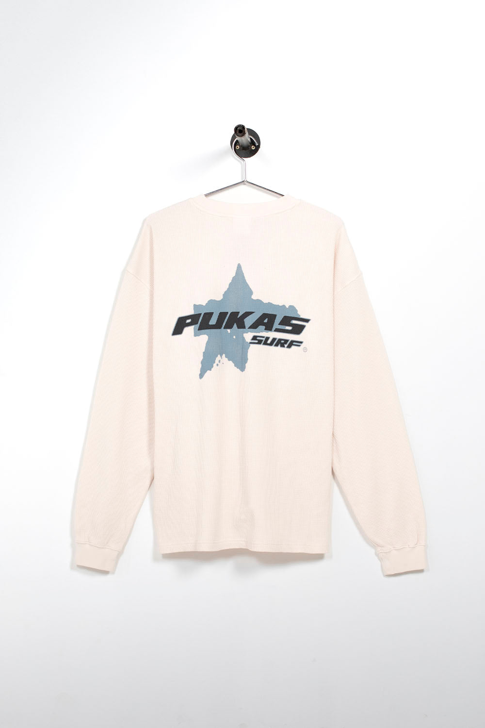A cream-colored long-sleeve t-shirt hanging on a black hanger against a white wall. The shirt features a large graphic on the chest with a gray star shape and the text 'PUKAS SURF' in black and gray.