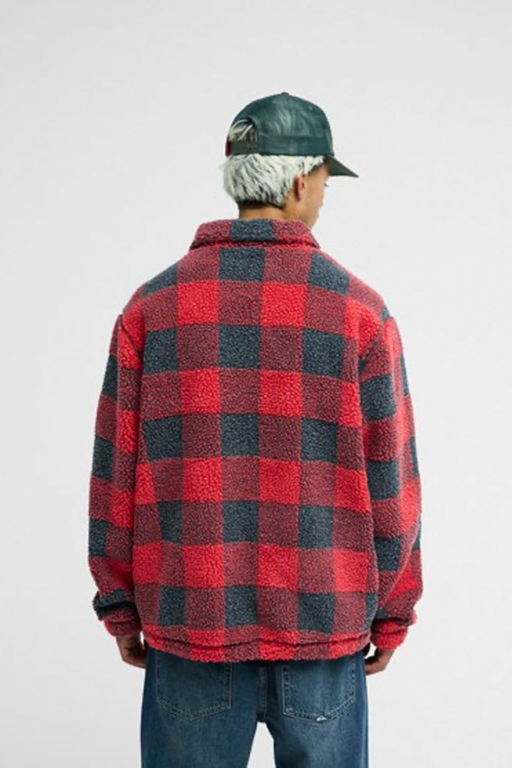 A person shown from the back wearing a red and dark gray buffalo plaid sherpa fleece jacket, blue jeans, and a dark green cap against a light gray studio background.