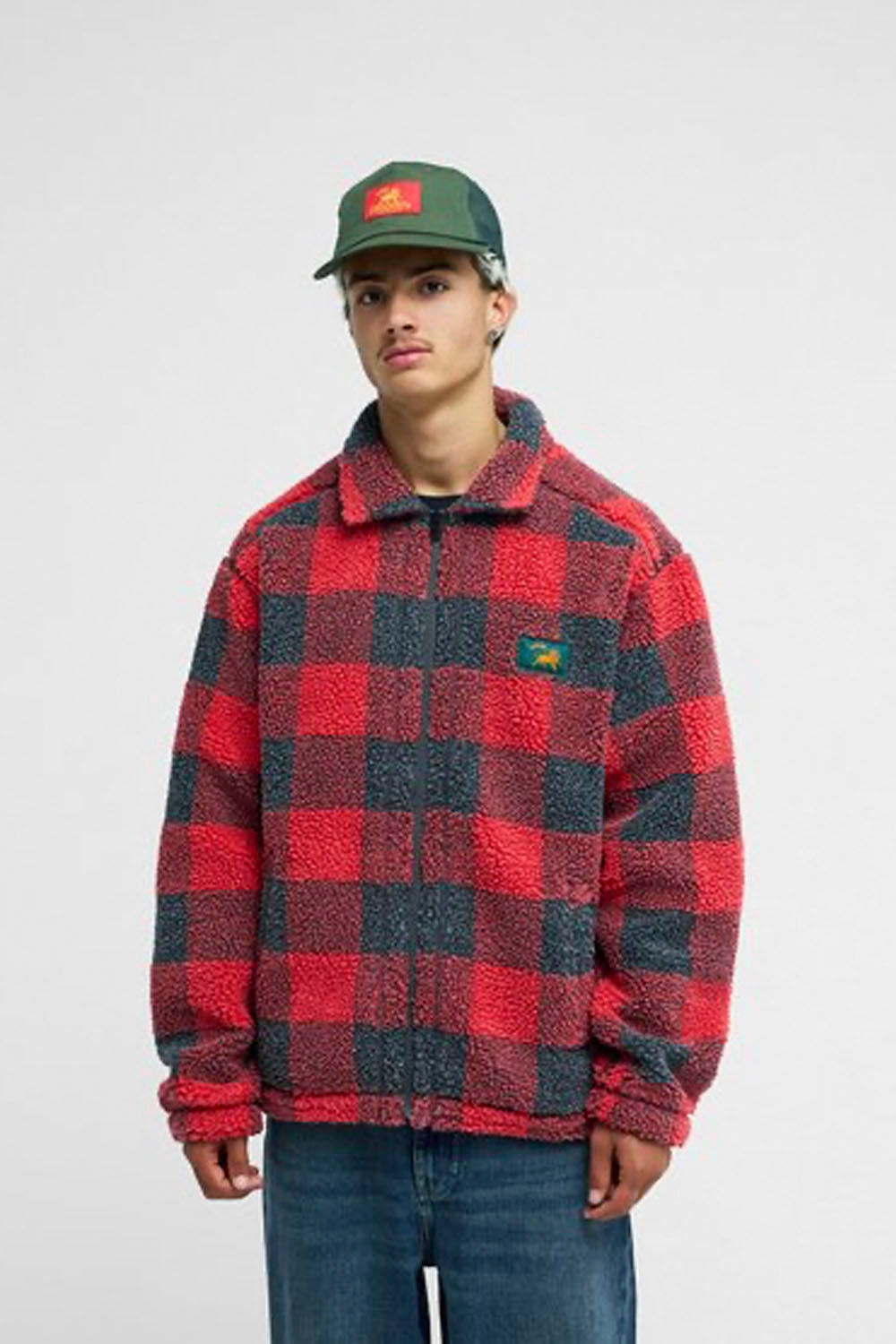 A model stands against a light gray studio background wearing a red and black buffalo plaid fleece jacket over a black shirt, blue jeans, and a green cap.