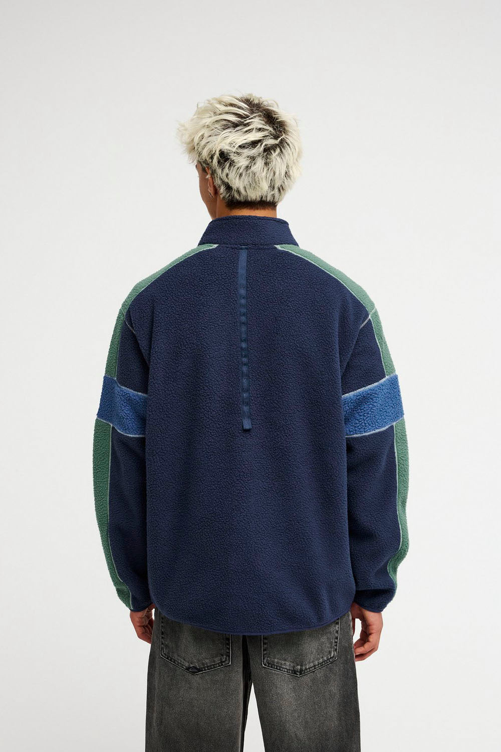 A person viewed from the back wearing a quarter-zip fleece pullover in navy blue with green side panels and blue stripe accents on the sleeves, paired with gray jeans, against a plain light gray studio background.