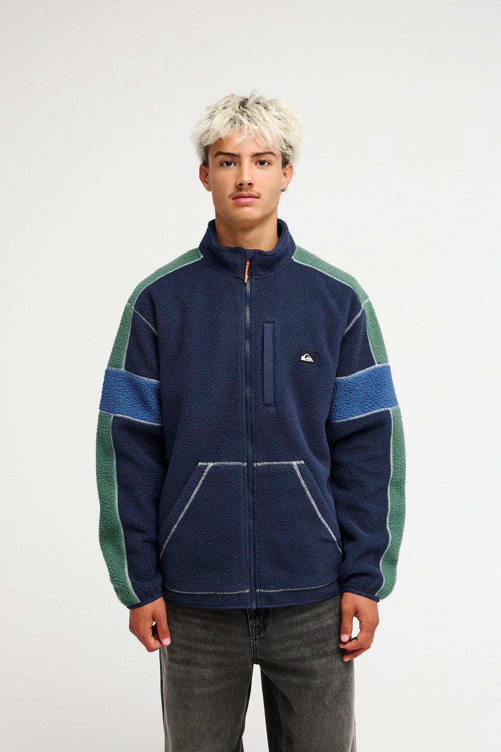 A model wearing a vintage Quiksilver fleece jacket in navy blue with green and light blue color-blocked panels on the shoulders and sleeves, paired with dark gray jeans, against a plain light gray background.