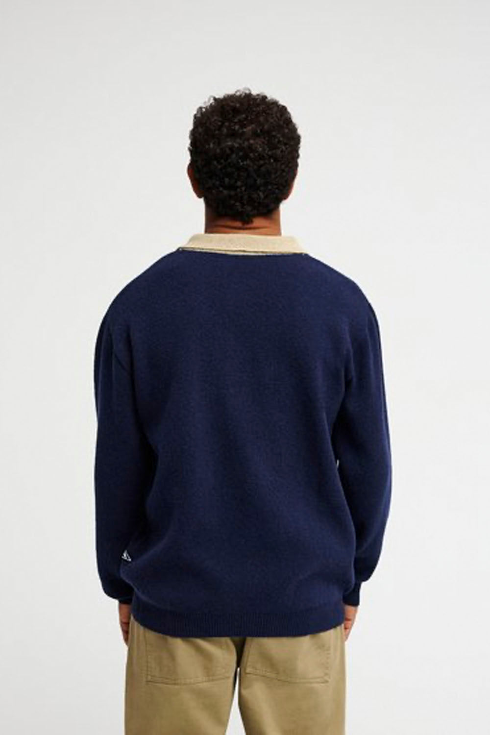 A man viewed from behind wearing a navy blue crewneck sweater with a beige collar, paired with khaki pants, against a plain light gray background.