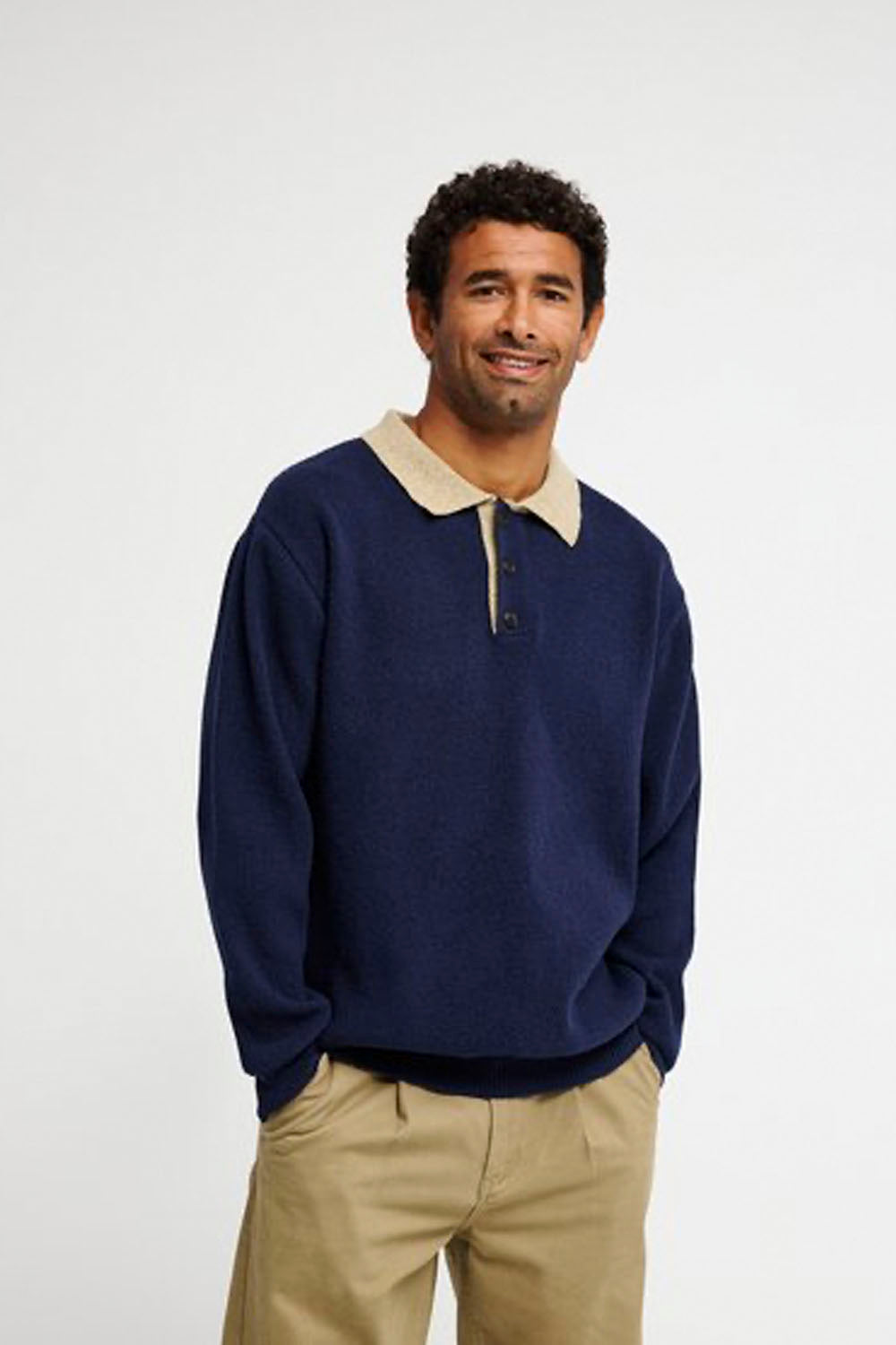 A man wearing a navy blue long-sleeve polo sweater with a beige collar, paired with khaki pants, standing against a light gray studio background.