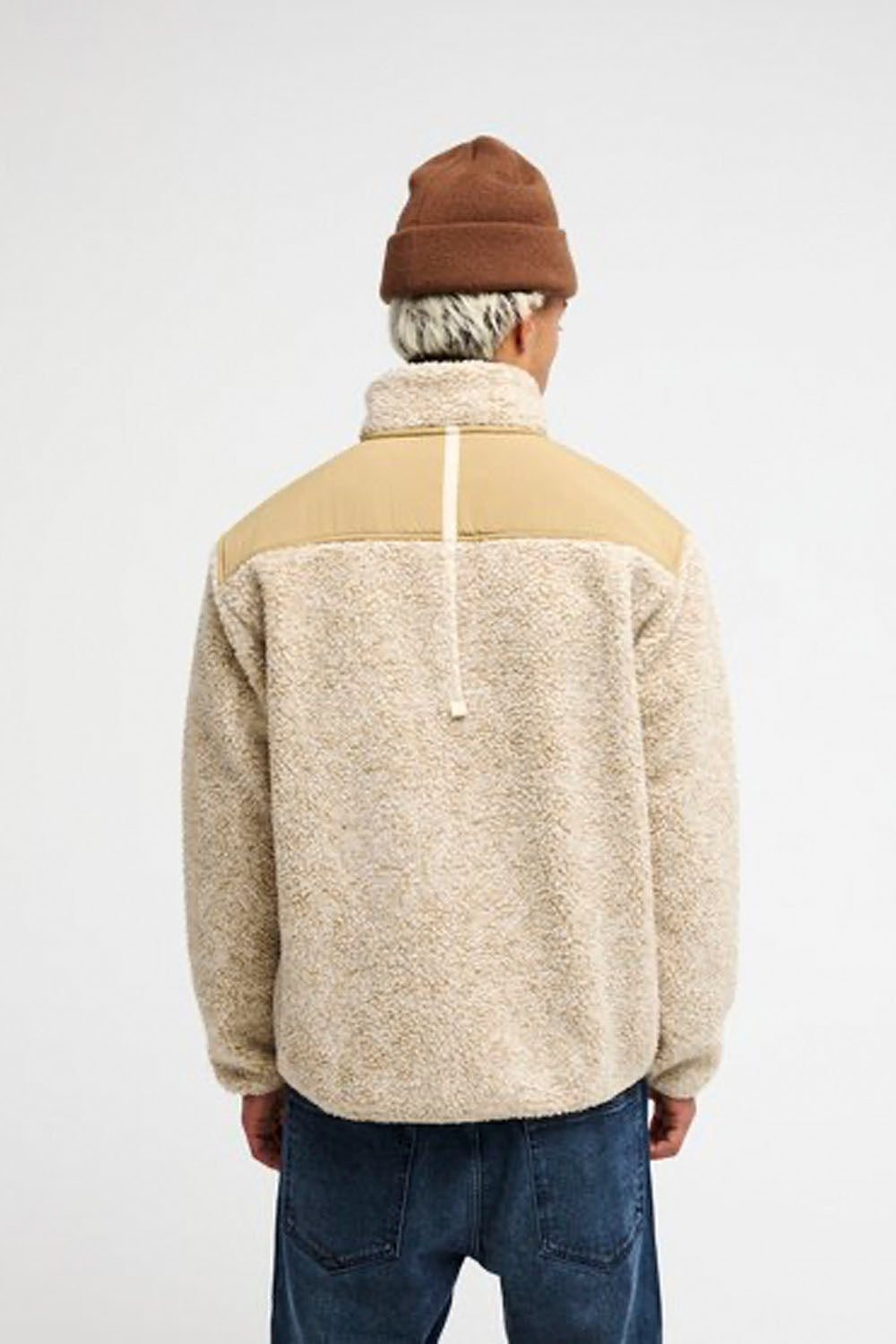 A person shown from the back wearing a sherpa fleece pullover jacket with a tan yoke and a brown knit beanie, against a plain light gray studio background.