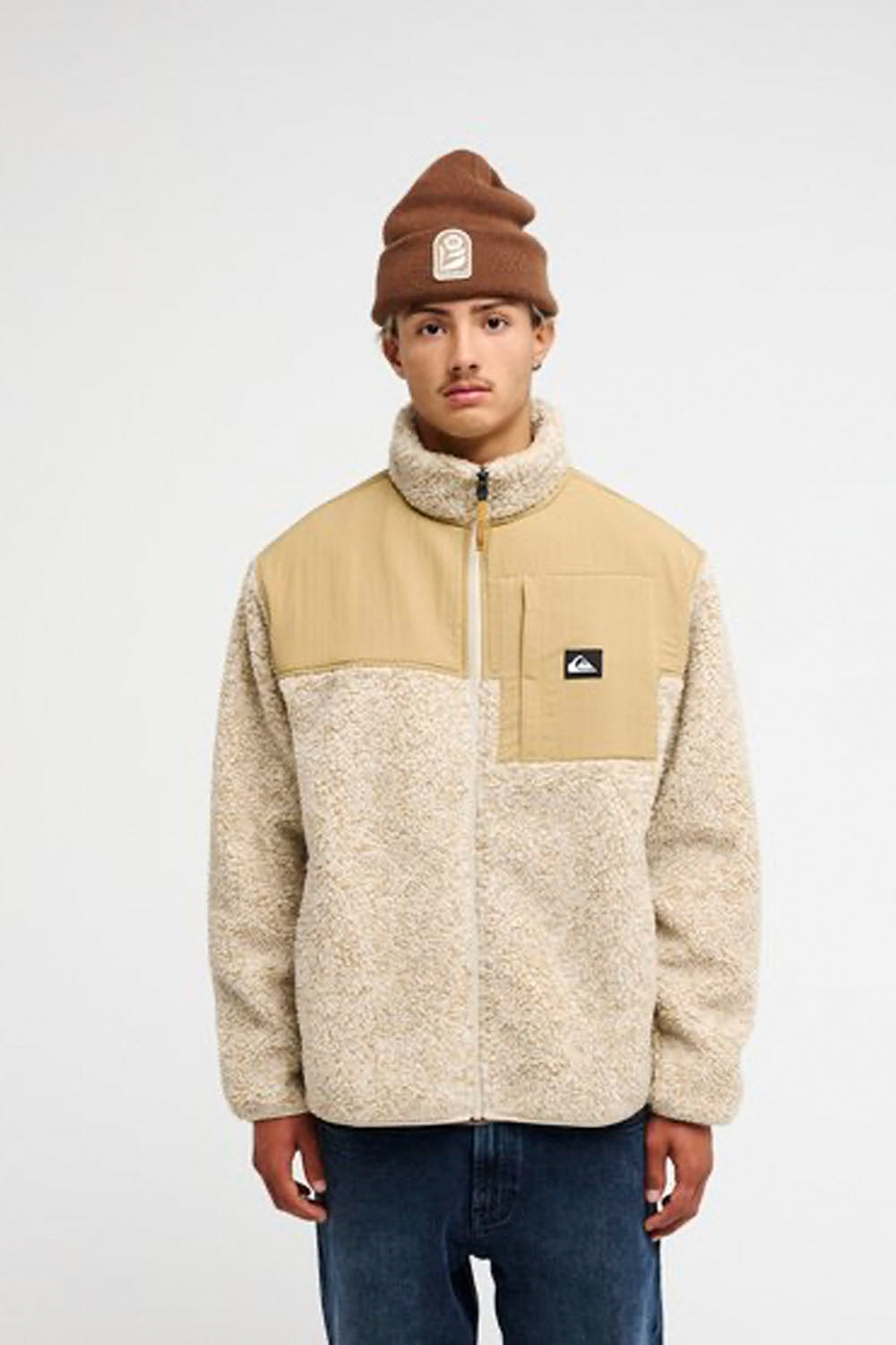 A model wearing a beige and cream color-blocked fleece jacket with a brown beanie, standing against a light gray studio background.