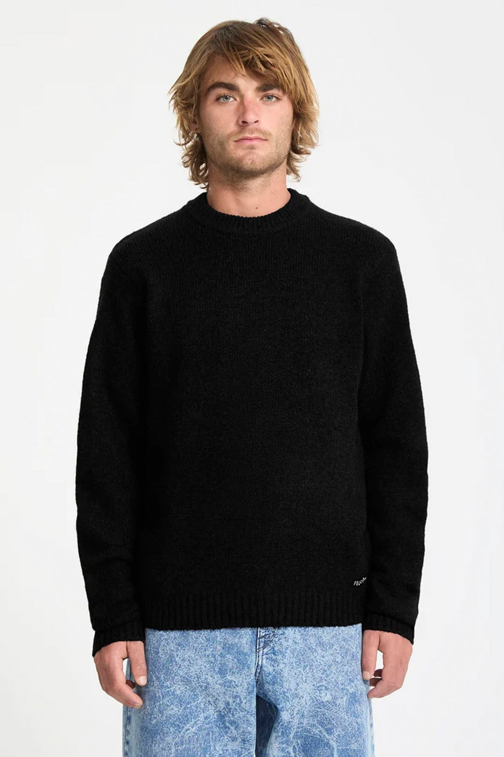 A man wearing a black knit crewneck sweater and light blue jeans, standing against a plain white studio background.