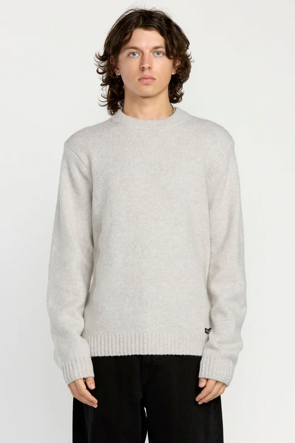 A man wearing a light gray knit crewneck sweater and black pants, standing against a plain white studio background.