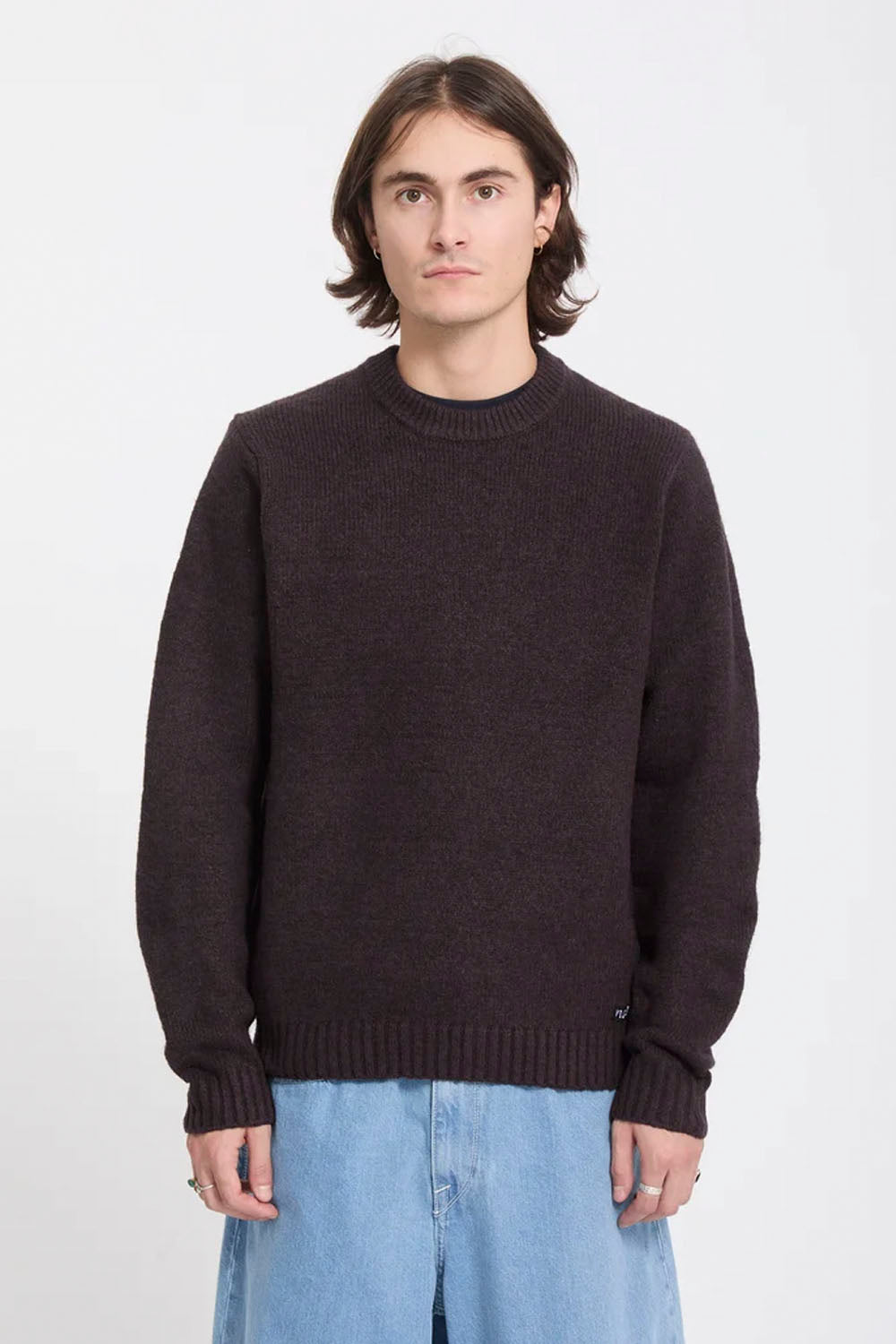 A man wearing a dark brown knit crewneck sweater and light blue jeans, standing against a plain white studio background.
