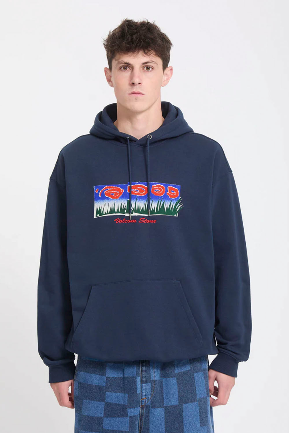 A man wearing a navy blue pullover hoodie with a colorful graphic print on the chest, paired with blue and gray checkered pants, against a plain white studio background.