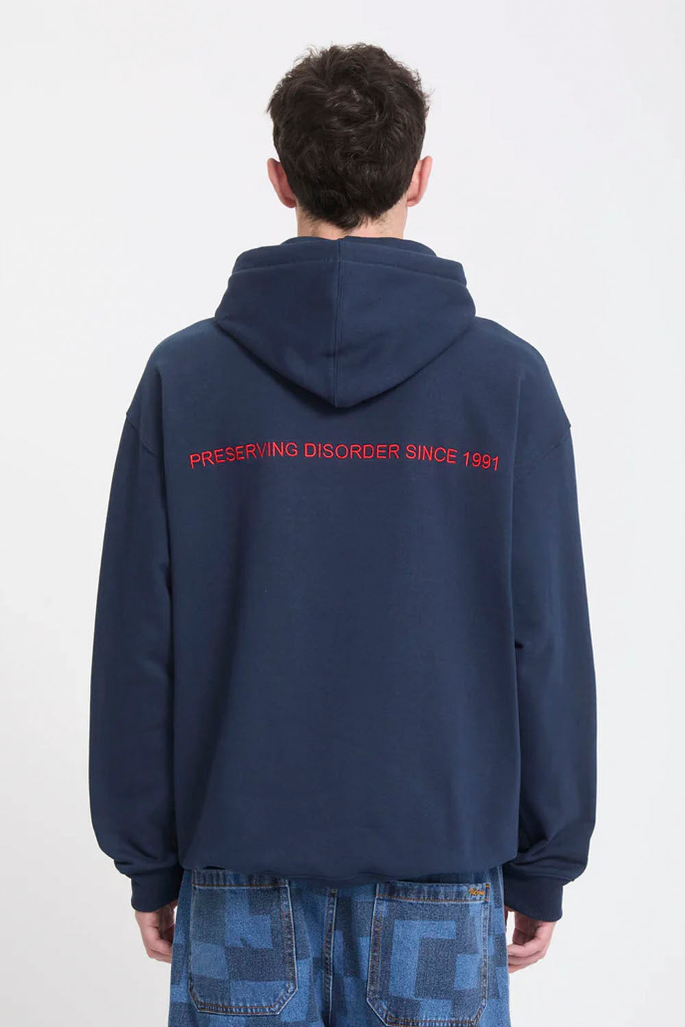 A person viewed from the back wearing a navy blue hoodie with red text reading 'PRESERVING DISORDER SINCE 1991' across the upper back, paired with blue jeans against a plain white background.