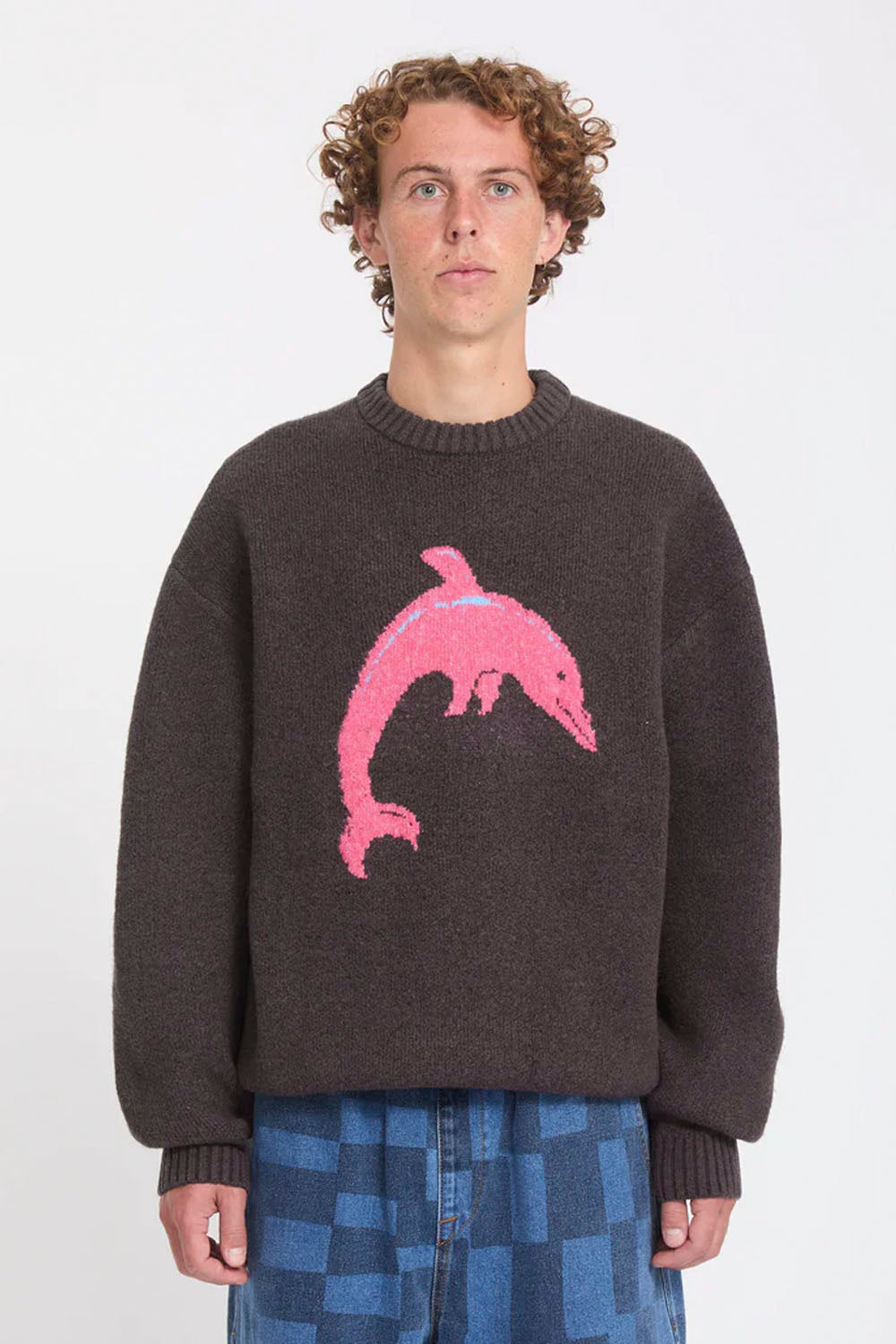 A model wearing a dark brown knit sweater with a large pink dolphin motif on the front, paired with blue checkered pants, against a plain light gray background.