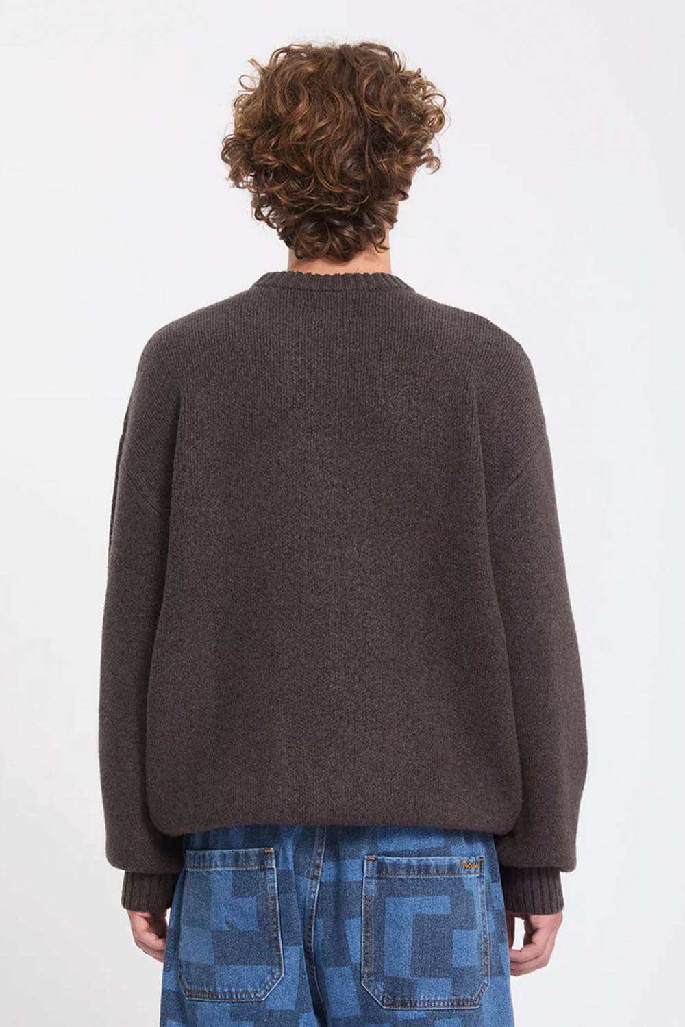 A person shown from the back wearing a dark brown knit sweater and blue patterned jeans against a plain light gray studio background.