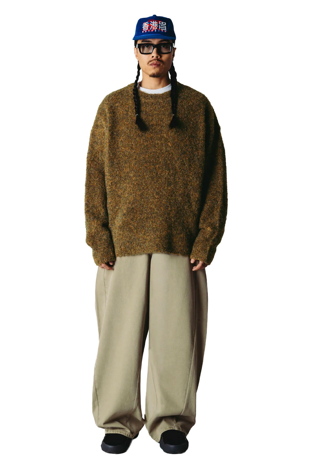 A person stands against a plain white background wearing an oversized brown knit sweater, loose light beige pants, black shoes, glasses, and a blue cap with Japanese text.