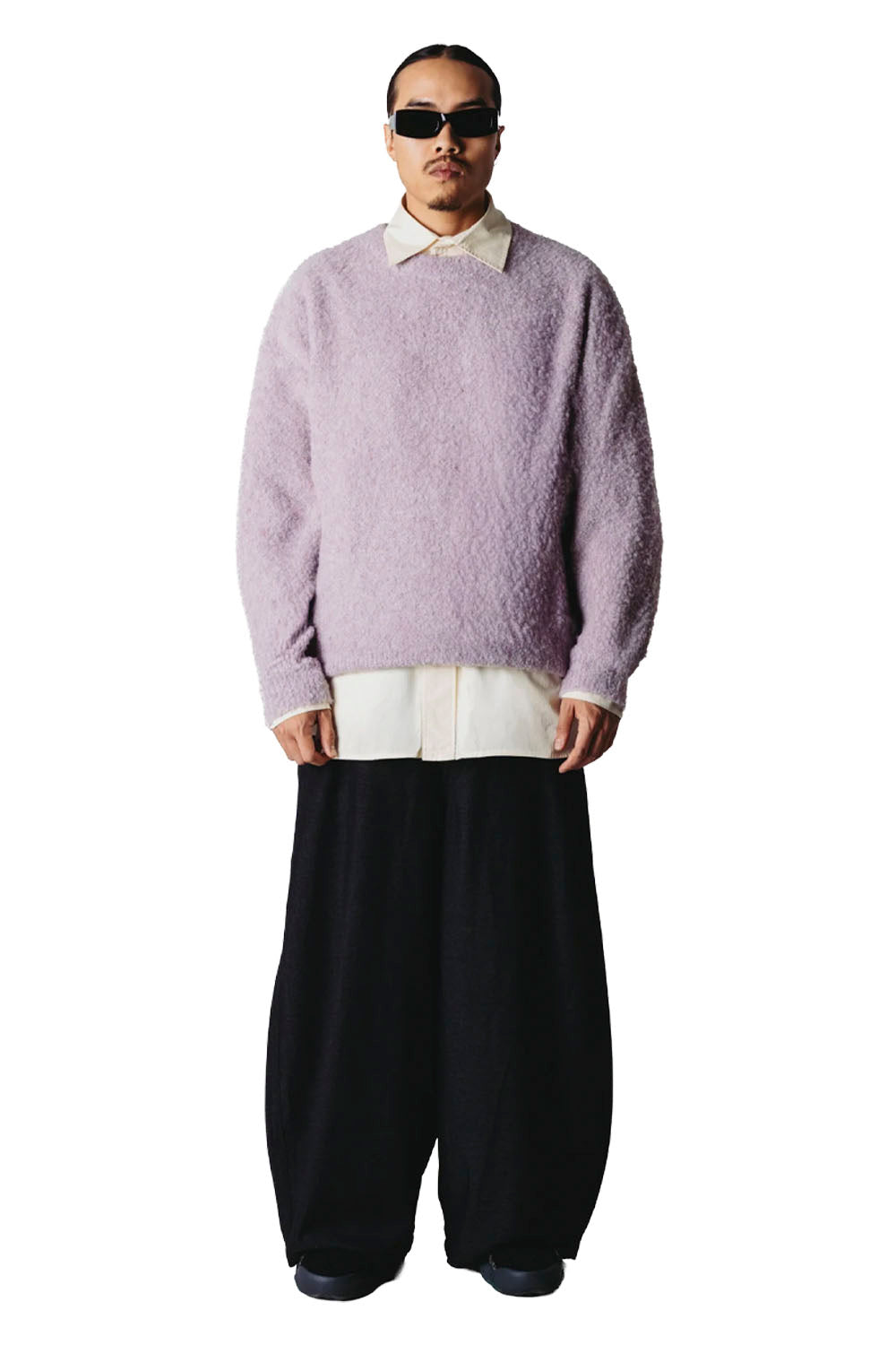 A person wearing a fuzzy light lavender cardigan over a cream shirt and loose black pants, photographed against a plain white studio background.