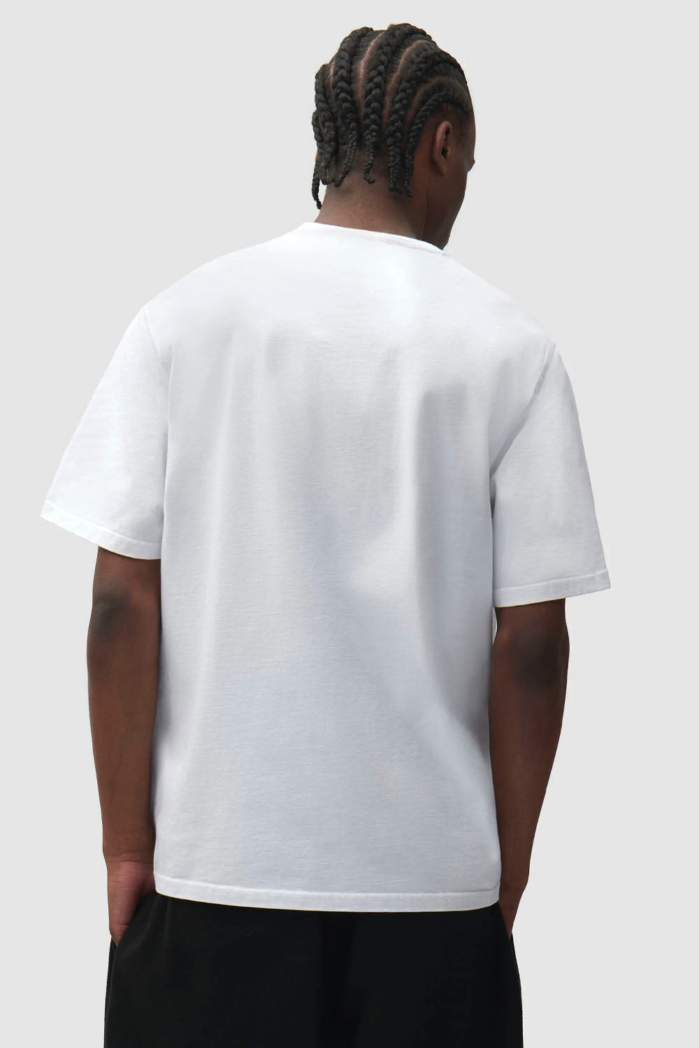 A man viewed from behind wearing a plain white short-sleeve crewneck t-shirt and black pants, set against a solid light gray background.