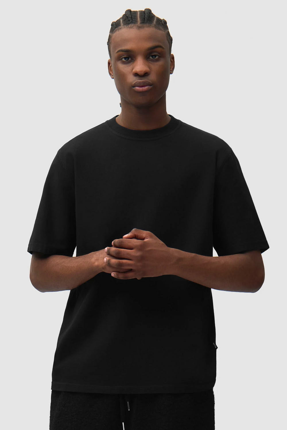 A man wearing a plain black short-sleeve crewneck t-shirt, standing against a light gray studio background with his hands clasped in front.