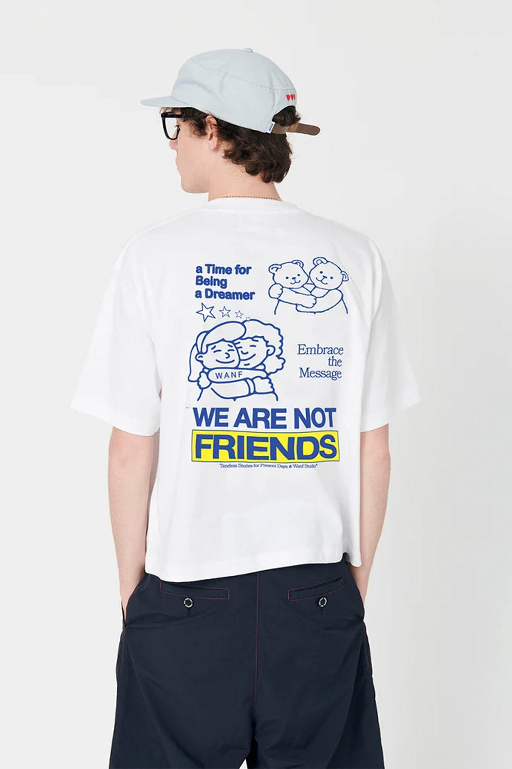 A model shown from the back wearing a white graphic t-shirt with blue and yellow text and illustrations that read 'WE ARE NOT FRIENDS'. The model is also wearing a light gray cap and dark navy pants against a clean white background.