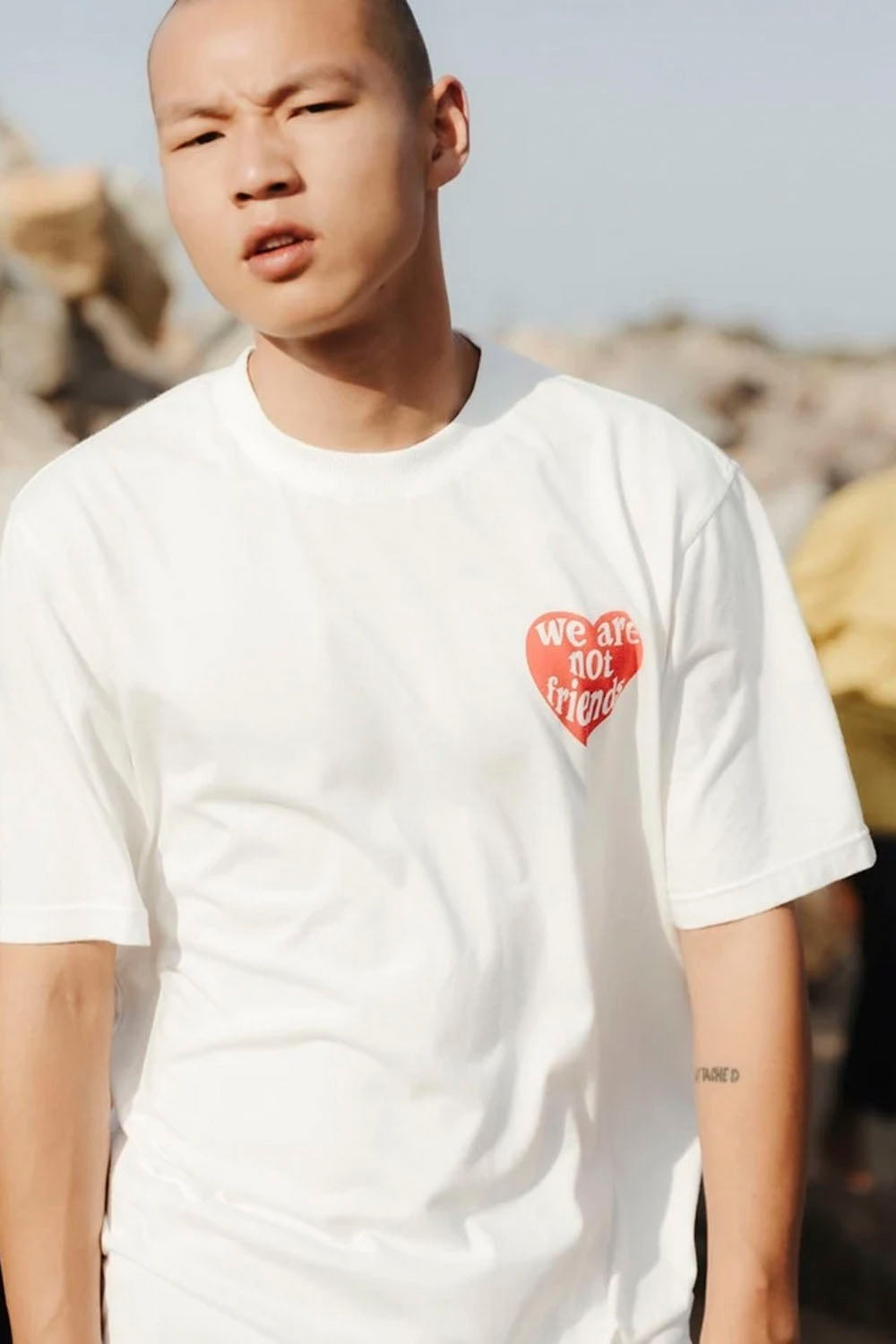 A man wearing a white crewneck t-shirt with a red heart graphic on the left chest that reads 'WE ARE NOT friends'. The background is a blurred outdoor rocky landscape.