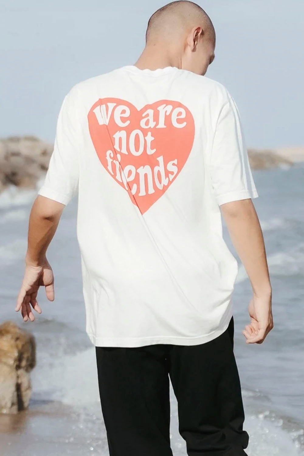 A man standing on a beach, viewed from behind, wearing a white t-shirt with a large coral-pink heart graphic on the back containing the text 'we are not friends' in white.