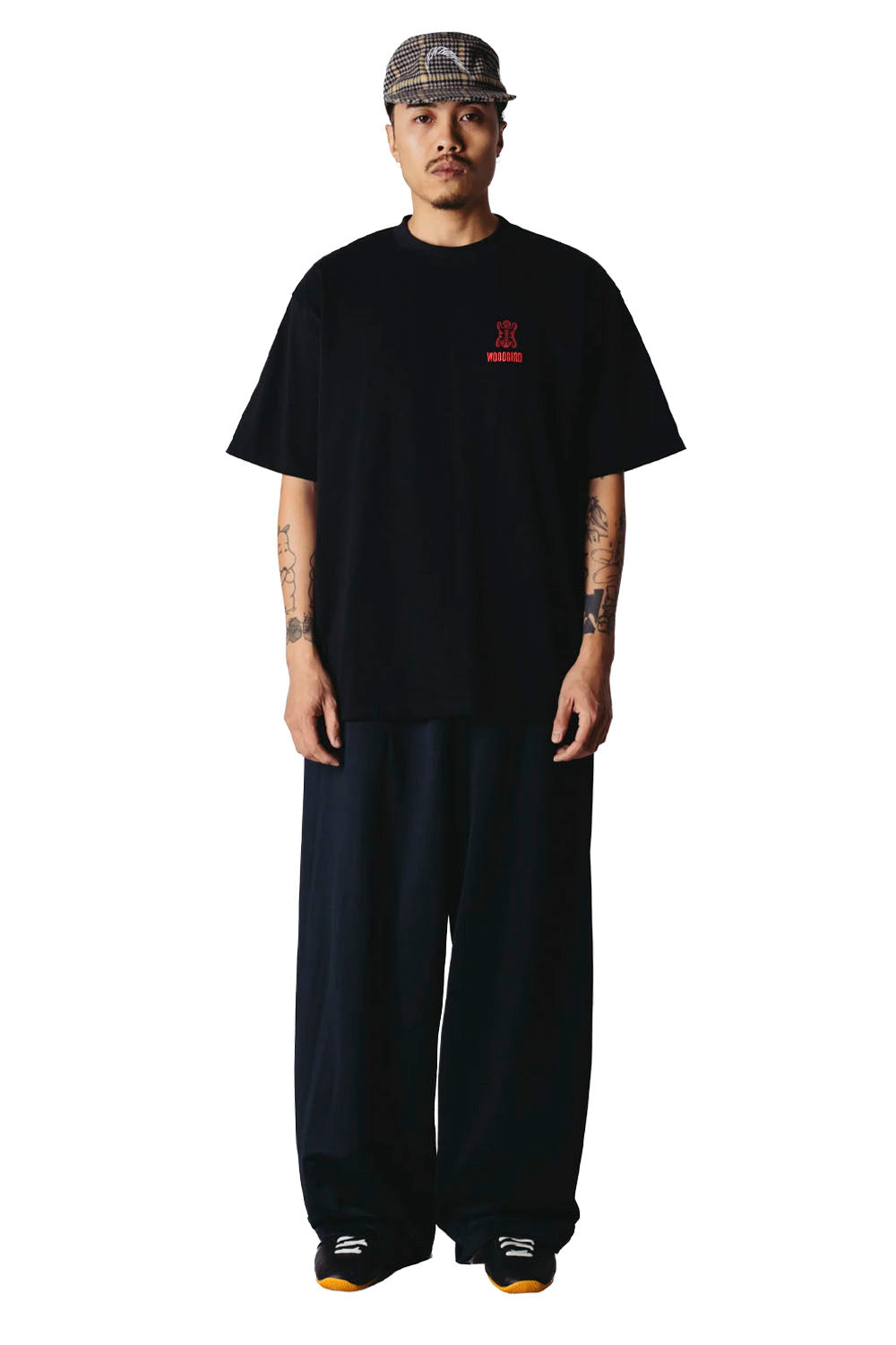 A man modeling a black oversized t-shirt with a small red graphic on the left chest, paired with matching black wide-leg pants, a patterned cap, and sneakers, shot against a solid white background.