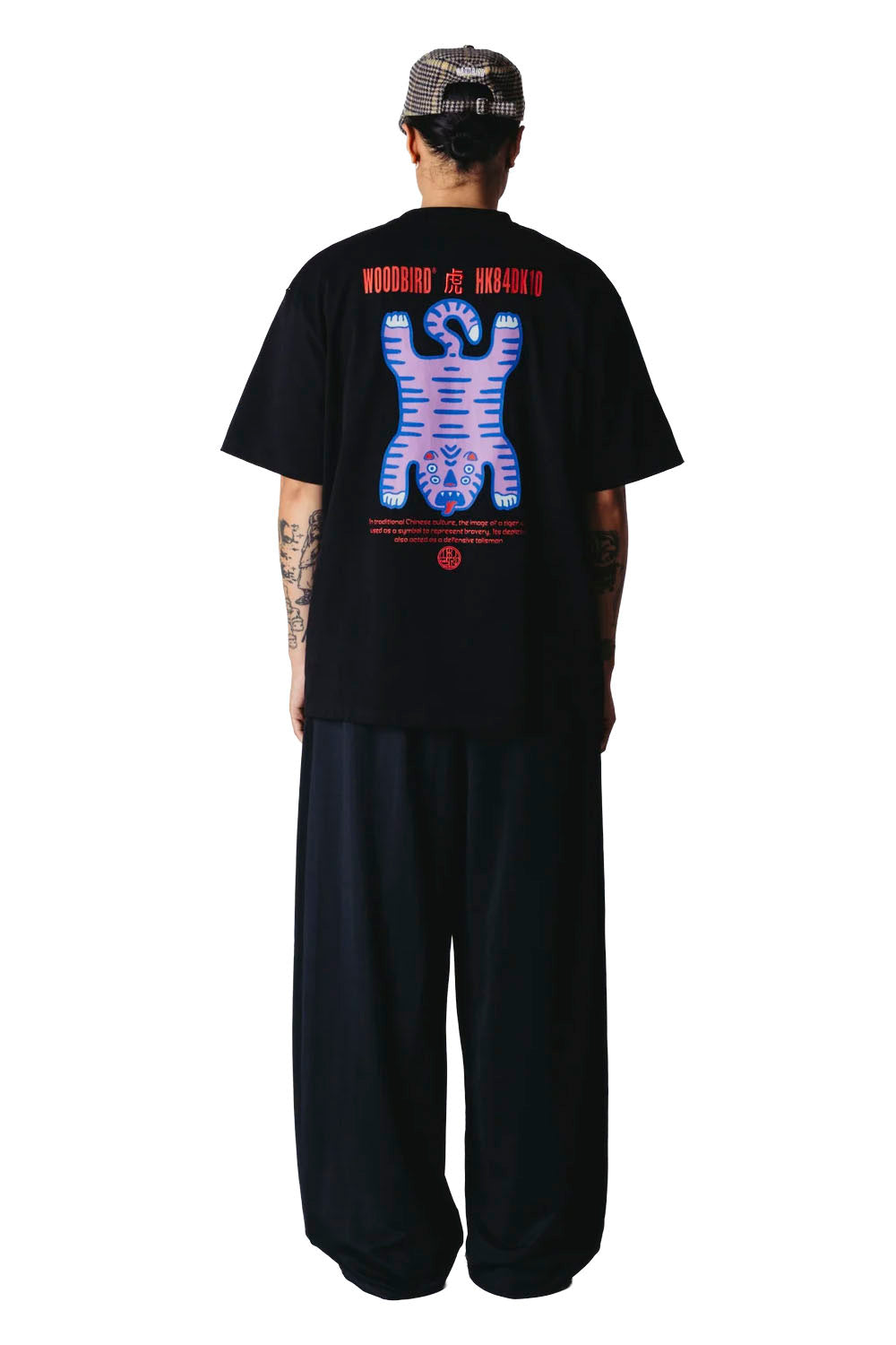 A man viewed from the back wearing a black t-shirt with a large colorful graphic print and text, paired with loose black pants against a white background.