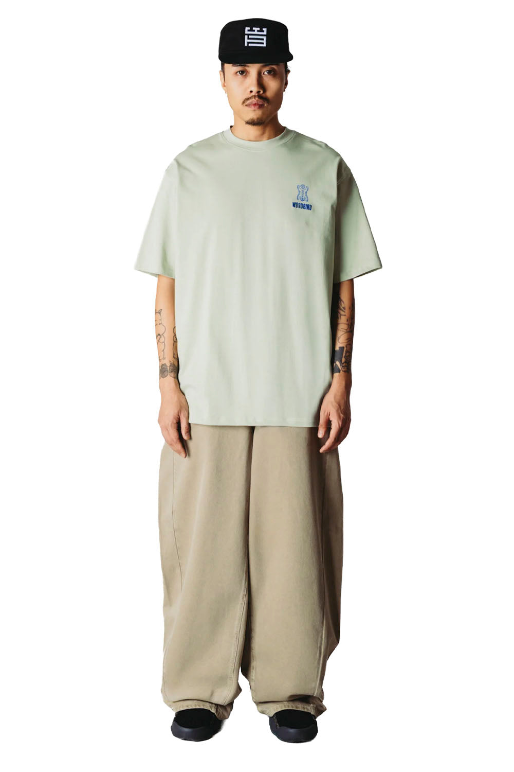 A model stands against a white background wearing a light sage green t-shirt with a small blue graphic on the left chest, paired with baggy beige pants and a black baseball cap.