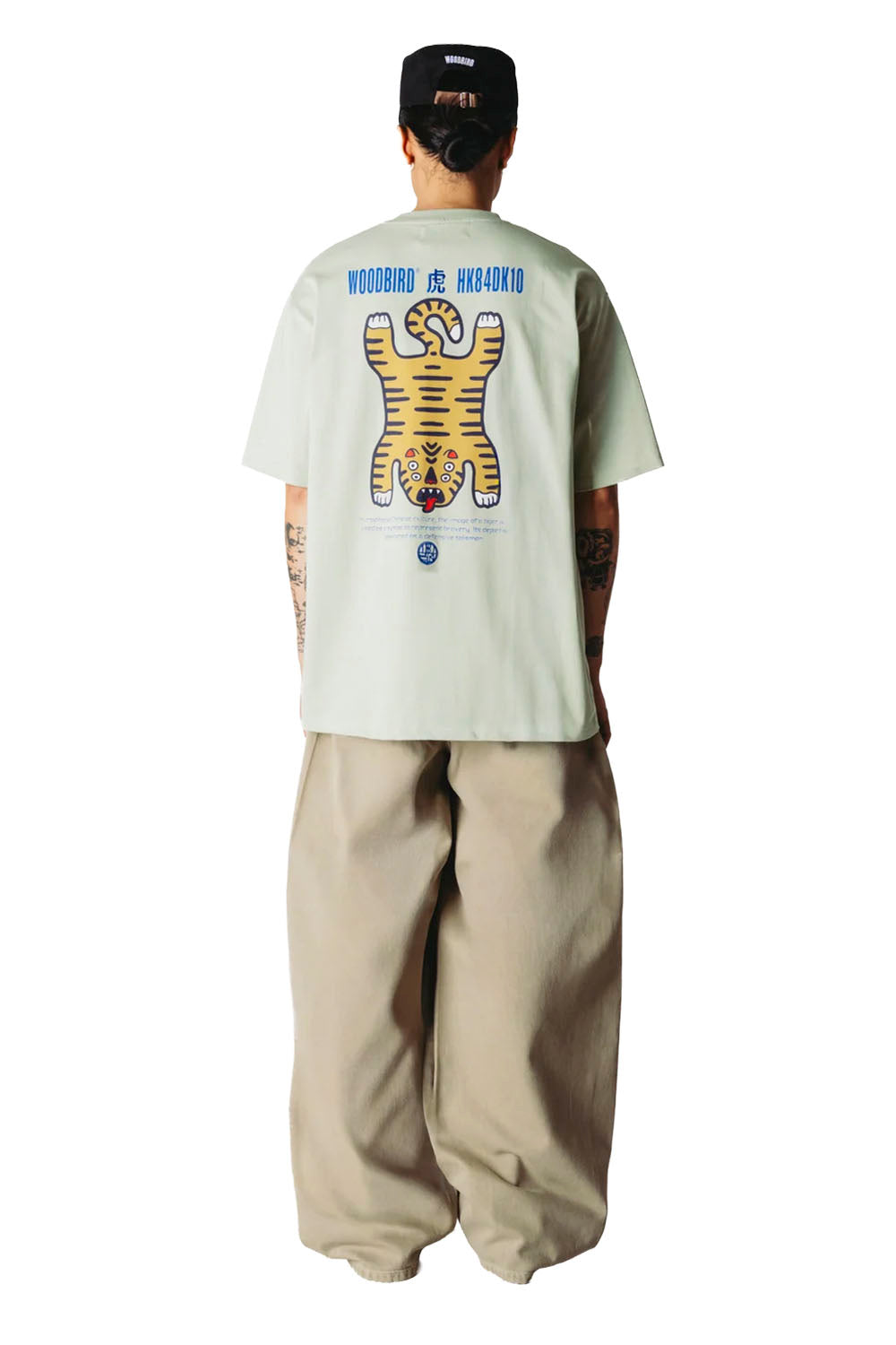 A person shown from the back wearing a light sage green t-shirt with a large yellow tiger graphic and blue text on the back, paired with beige baggy pants and a black cap.
