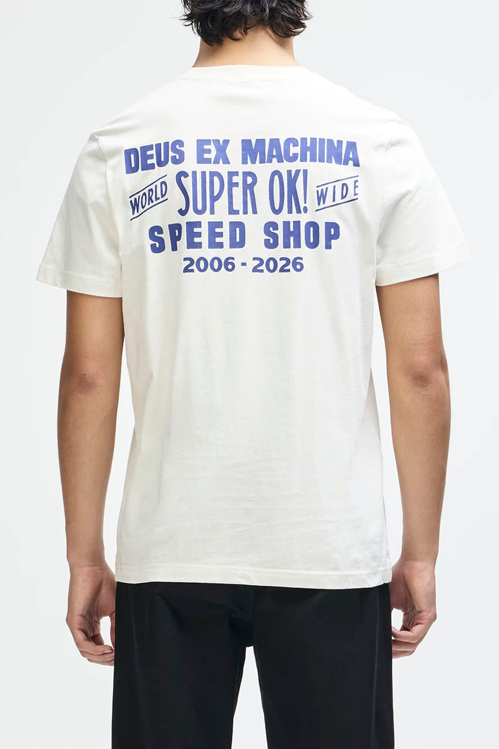 A man viewed from the back wearing a white t-shirt with a blue graphic print that reads 'DEUS EX MACHINA WORLD SUPER OK! WIDE SPEED SHOP 2006 - 2026'.