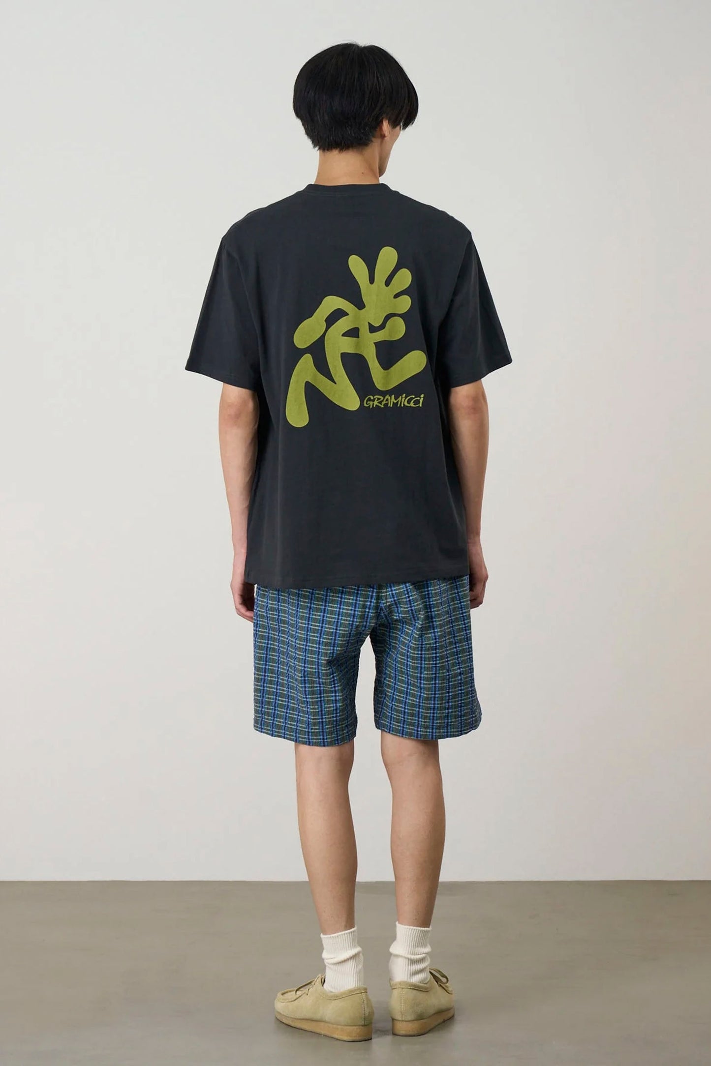 Person wearing a Black Gramicci Running Man Tee with yellow graphics and plaid shorts on a plain background