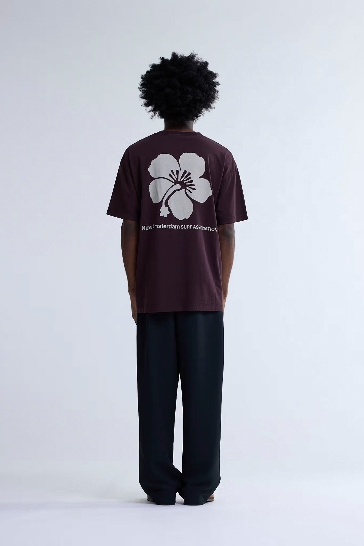 Person wearing a brown t-shirt with a white flower design on the back, standing against a white background.