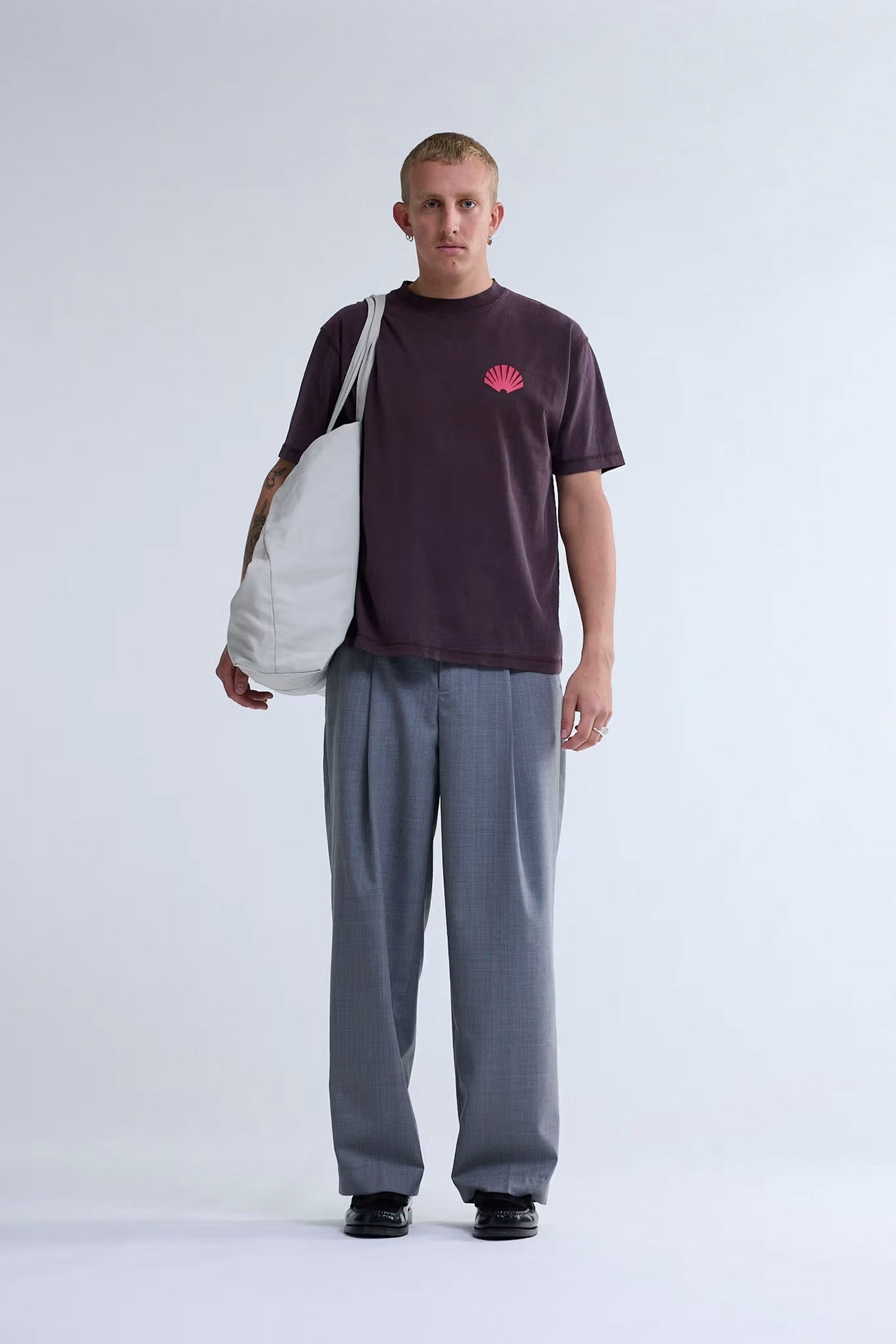 Man wearing a dark t-shirt with a logo and gray pants, holding a white bag on a light gray background