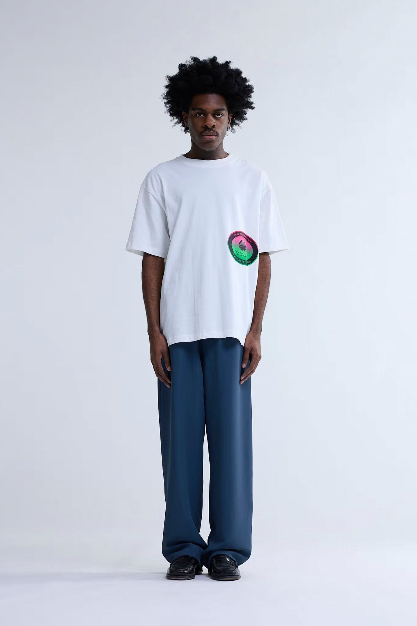 Person wearing New Amsterdam Surf Association Surf Centre Tee in Washed White with a colorful design and blue pants on a white background