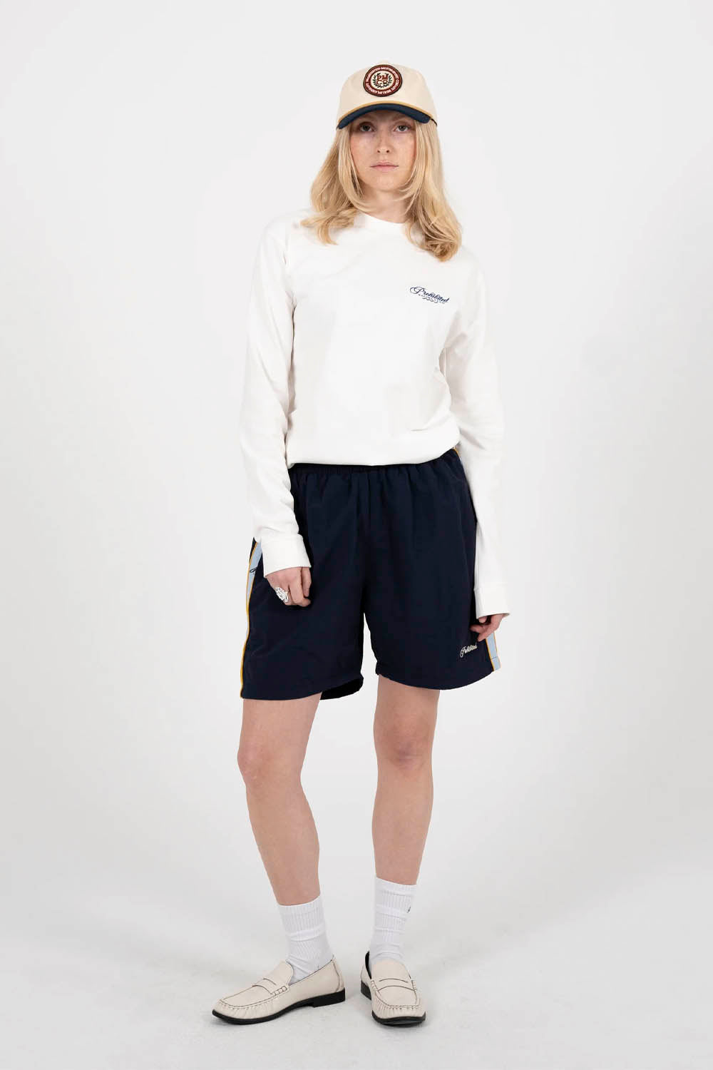 Person wearing a white sweatshirt, navy shorts, and a cap on a white background
