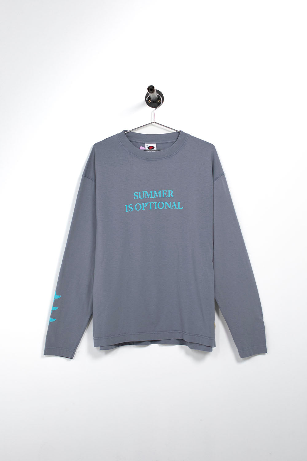 A gray long-sleeve t-shirt hanging on a wall hook, featuring the text 'SUMMER IS OPTIONAL' printed in light blue on the chest.