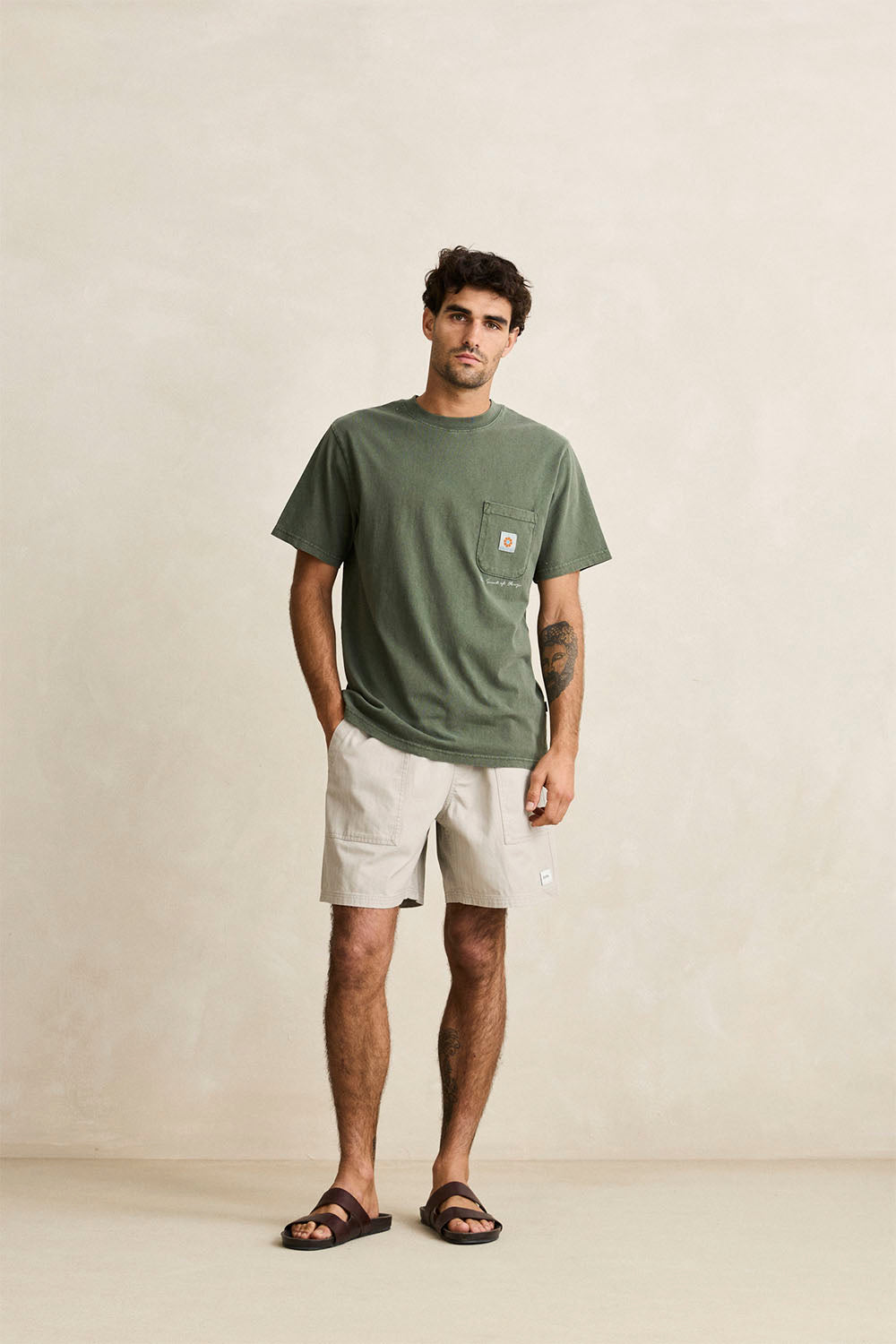 A man wearing an olive green Rhythm pocket t-shirt, off-white shorts, and black sandals, standing against a textured beige wall.