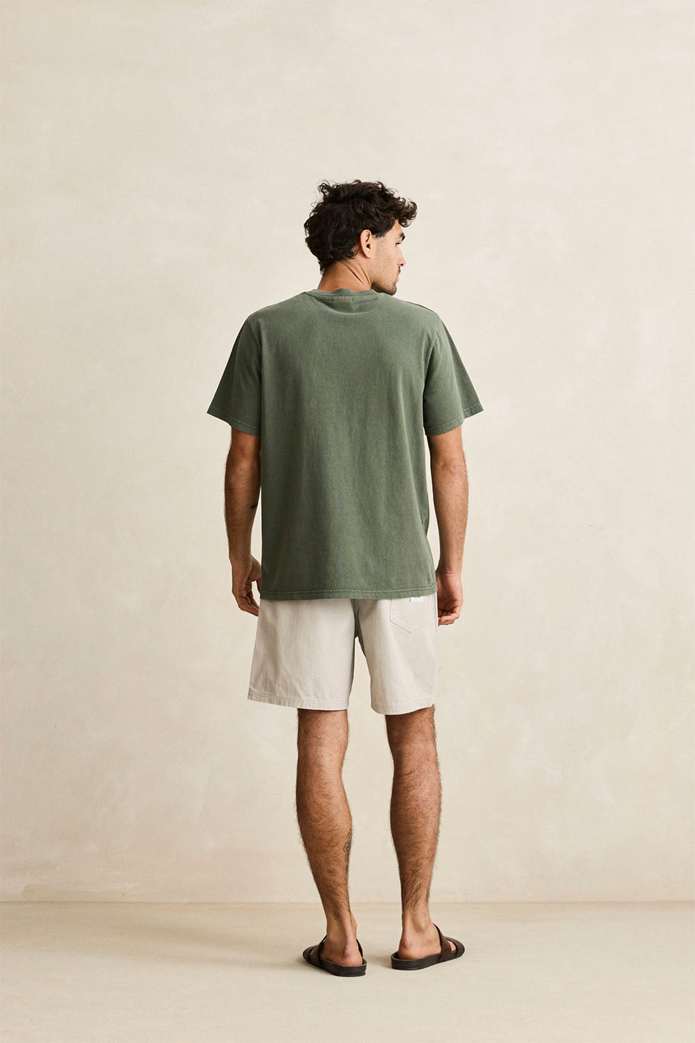 A man is shown from behind wearing an olive green short-sleeve t-shirt and off-white shorts, standing against a neutral beige textured wall.