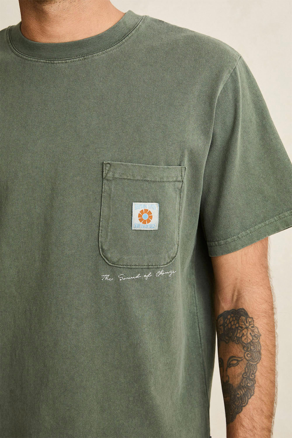 A close-up of a person wearing an olive green short-sleeve pocket t-shirt with a small white chest patch featuring a colorful sun design and the text 'The Second of June' printed below it.