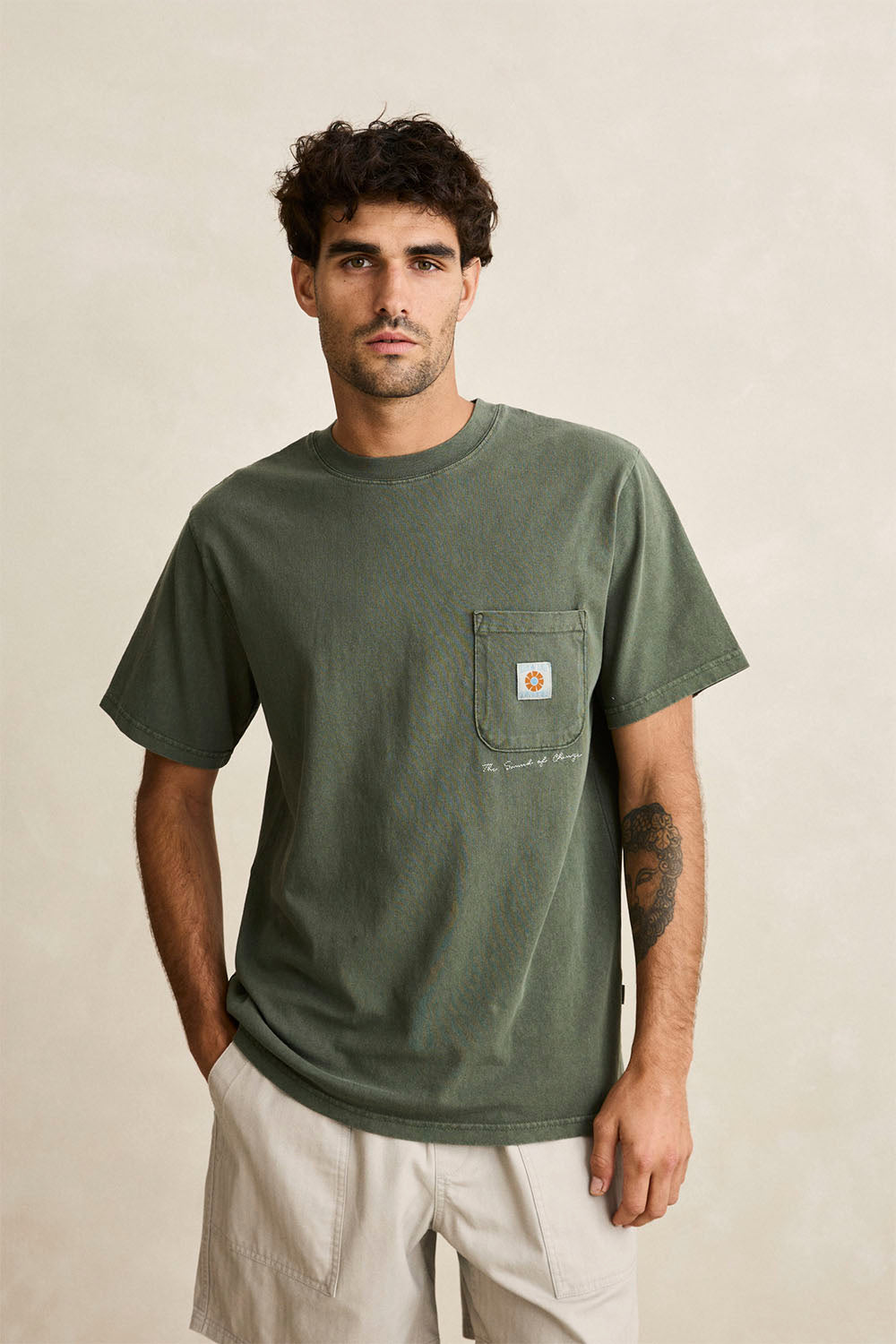 A man wearing an olive green Rhythm pocket t-shirt, off-white shorts, and black sandals, standing against a textured beige wall.